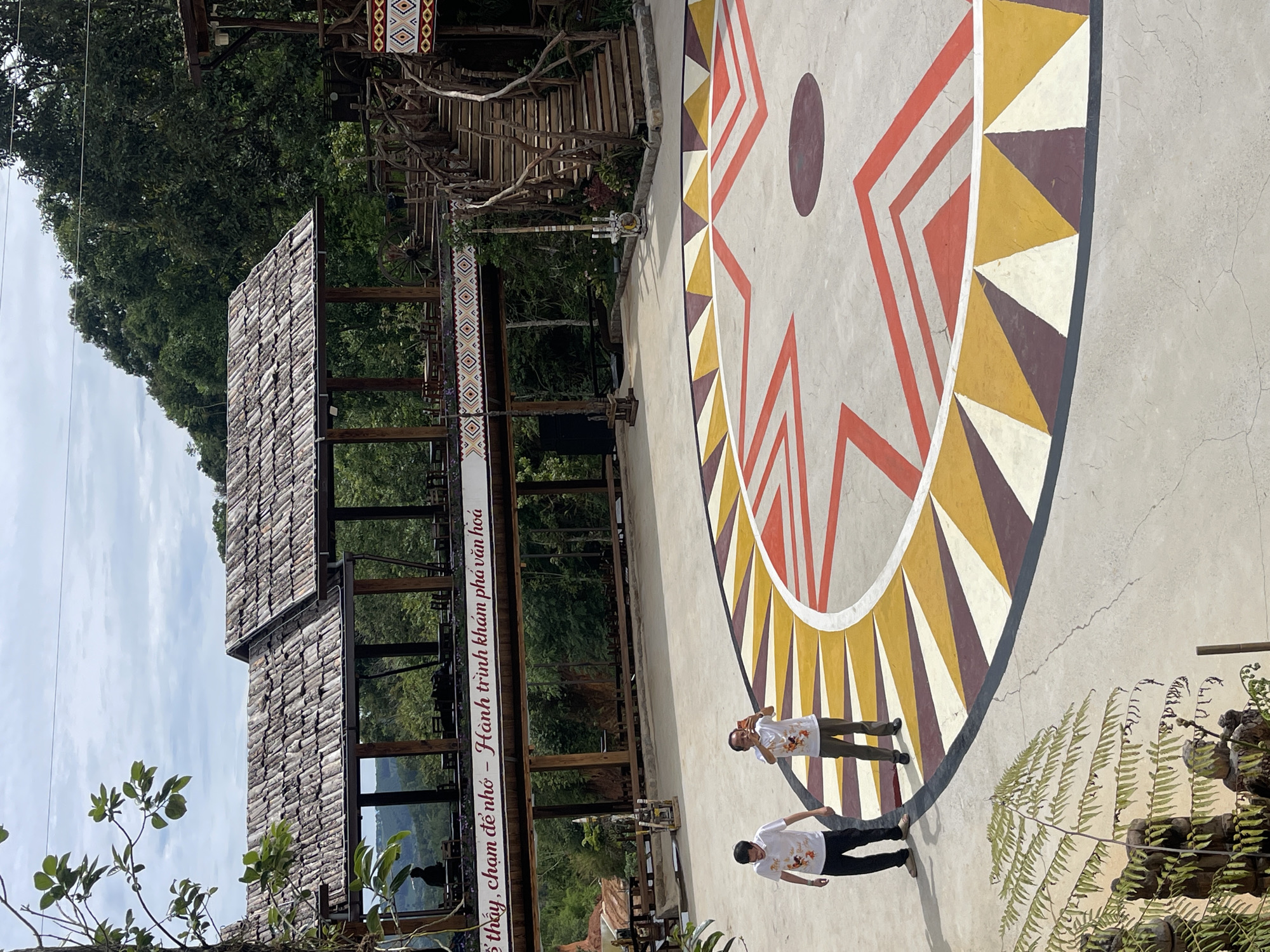The scene shows an outdoor space in Thôn 5, Vietnam, where a large circular design is painted on the ground. This design consists of concentric geometric shapes in colors of red, yellow, white, and brown. Two individuals are standing on the painted area. They are wearing white shirts with a design on them. Adjacent to the painted area is a structure with wooden beams and a tiled roof. A sign with Vietnamese text is affixed to the structure. The background includes greenery such as trees and ferns, indicating a natural setting.