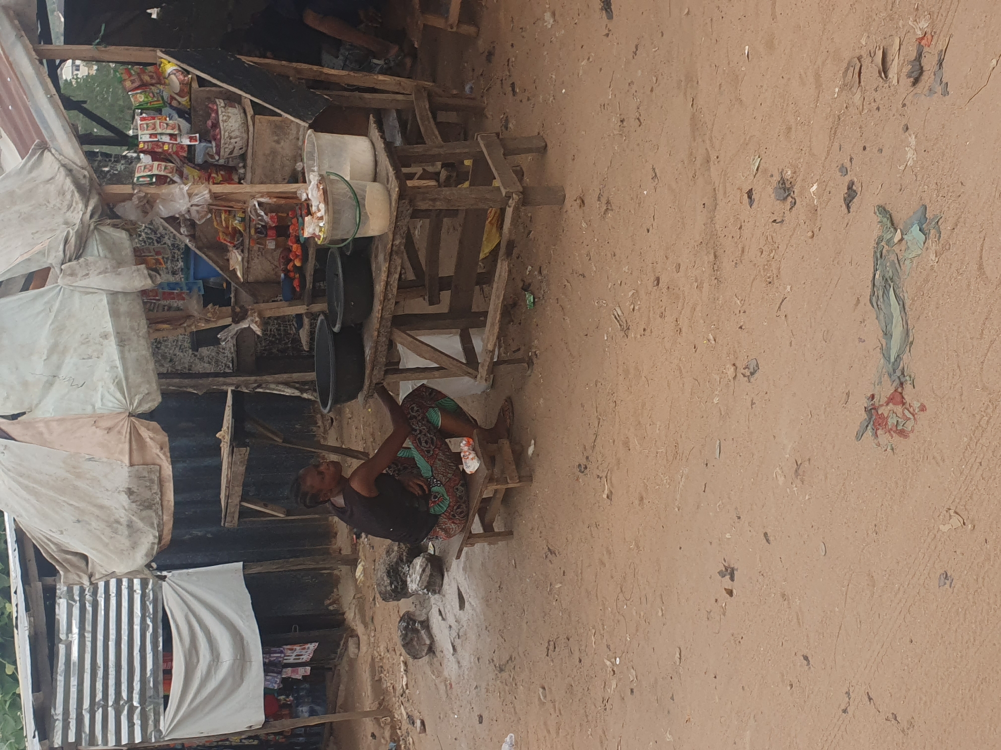 A woman sits on a small wooden stool next to a roadside kiosk in Lokoja, Nigeria. The kiosk, constructed from rough-hewn wood and corrugated metal, is stocked with packaged goods. Plastic containers and black basins sit on the kiosk's shelves. The ground is bare dirt. Litter is scattered on the ground.