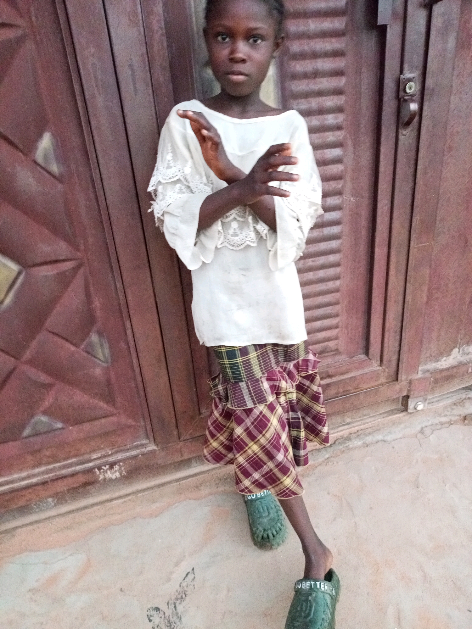 In Tempure, Nigeria, a young Black girl stands in front of a wooden door. She is wearing a white, lace-trimmed shirt and a plaid skirt. Her arms are crossed over her chest. She has on green clogs. The ground is sandy and there is a small metallic object on the door hinge.