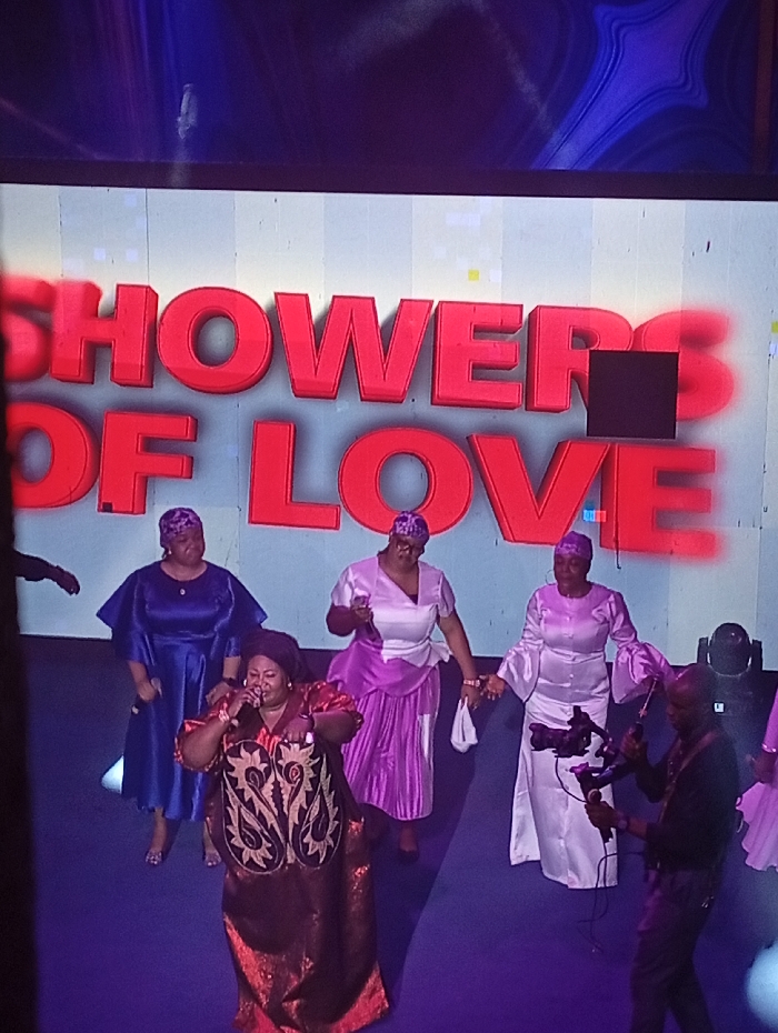 A group of five women are on stage with "Showers of Love" displayed behind them. The woman in the foreground wears a red and gold dress and sings into a microphone. The other women are dressed in purple, white, and blue dresses and hats. A man with a video camera is filming the women. The stage is blue, and the background is white with red lettering.