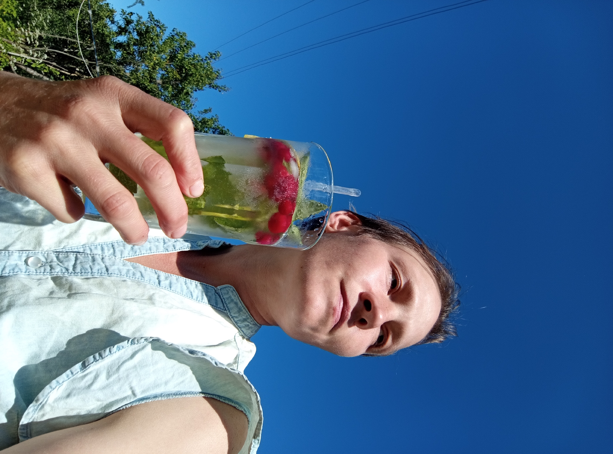 The media file shows a person holding a glass of iced beverage outdoors against a clear blue sky. The person, who appears to be female, is wearing a light blue denim shirt. The glass contains ice, mint leaves, raspberries, and a slice of lemon or lime. A clear plastic straw is visible in the glass. The hand holding the glass is positioned in the upper left of the frame. In the background, out of focus, are green trees and power lines. The image is taken from a low angle, looking upwards. The bright sunlight suggests it is daytime, likely a warm and sunny day. The overall mood seems relaxed and summery. No text is visible in the image. The location context is Ostrovtsy, Russia.