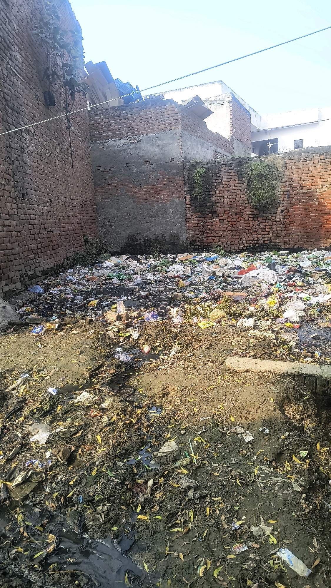 The image shows a large pile of garbage accumulated in an outdoor area within the city of Loni, India. The garbage consists of a vast amount of plastic waste, including bags, containers, and wrappers, mixed with organic debris such as leaves and mud. The area appears to be an open space enclosed by brick walls, some of which show signs of disrepair. There are no people visible in the image. The environment is unsanitary and suggests a lack of waste management. The overall scene depicts a significant litter problem.