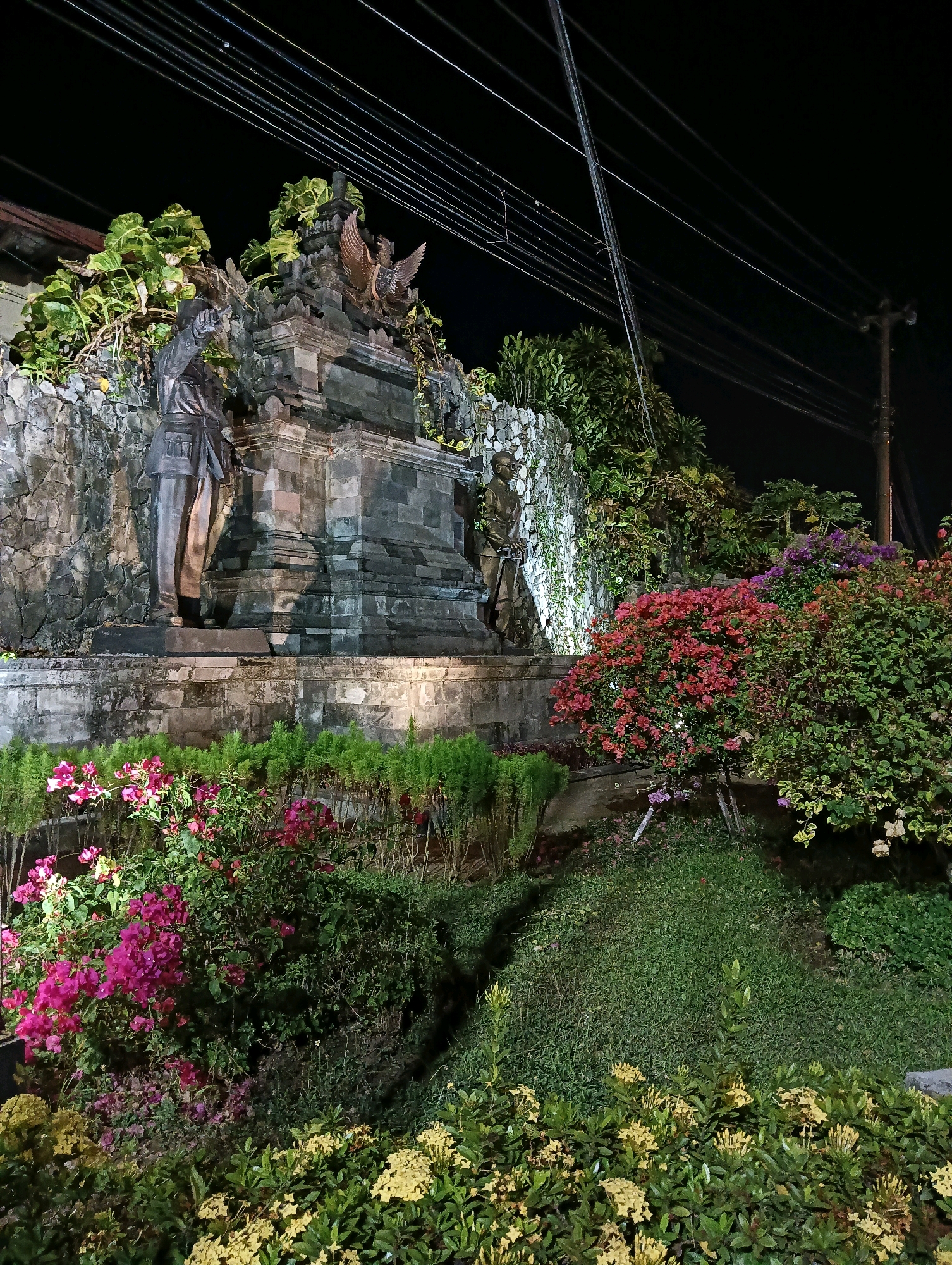 This image depicts a monument at night, located in Purworejo, Indonesia. The monument is constructed of stone and features two statues of men, one on the left and one on the right. A bronze eagle statue is positioned at the apex of the monument. The foreground is adorned with lush greenery and vibrant flowers in shades of pink, red, and yellow. Overhead, power lines stretch across the dark sky, with a utility pole visible on the right side of the frame. The lighting suggests artificial illumination of the monument and surrounding vegetation, creating a dramatic nighttime scene.