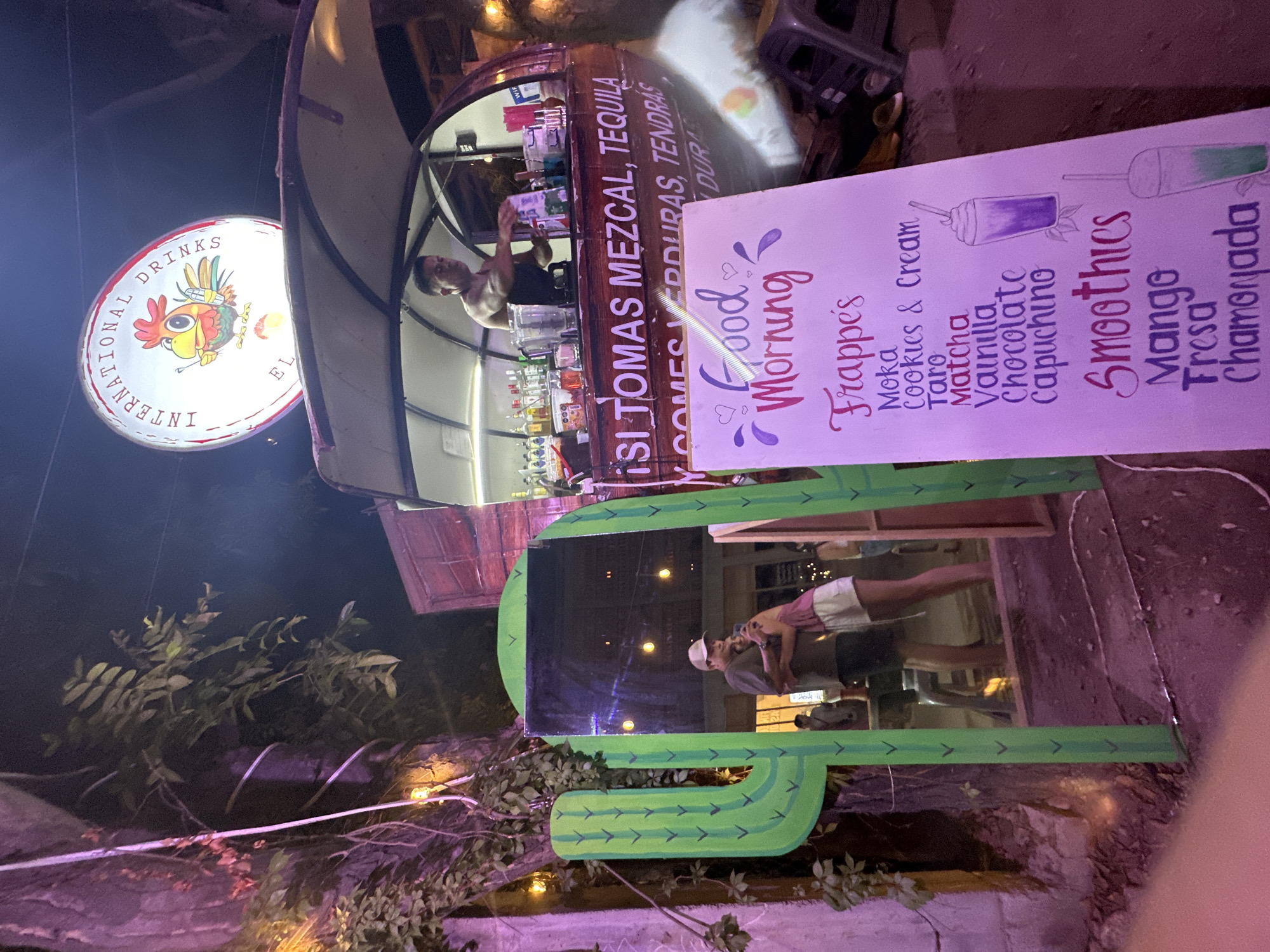 The image depicts a nighttime scene in what appears to be Brisas de Zicatela, Mexico, showcasing a mobile beverage cart. The cart is adorned with the words "ISI TOMAS MEZCAL, TEQUILA." A man stands inside the cart, seemingly preparing drinks. A sign next to the cart advertises "Good Morning" beverages, listing options like Frappe's (Moka, Cookies & Cream, Taro, Matcha, Vanilla, Chocolate, Capuchino) and Smoothies (Mango, Fresa, Chamonada). A cactus-shaped photo frame is positioned nearby, reflecting an image of people in the frame. An illuminated sign reading "International Drinks EL" is visible above the cart. The surrounding environment includes plants and what appears to be a dimly lit outdoor area.