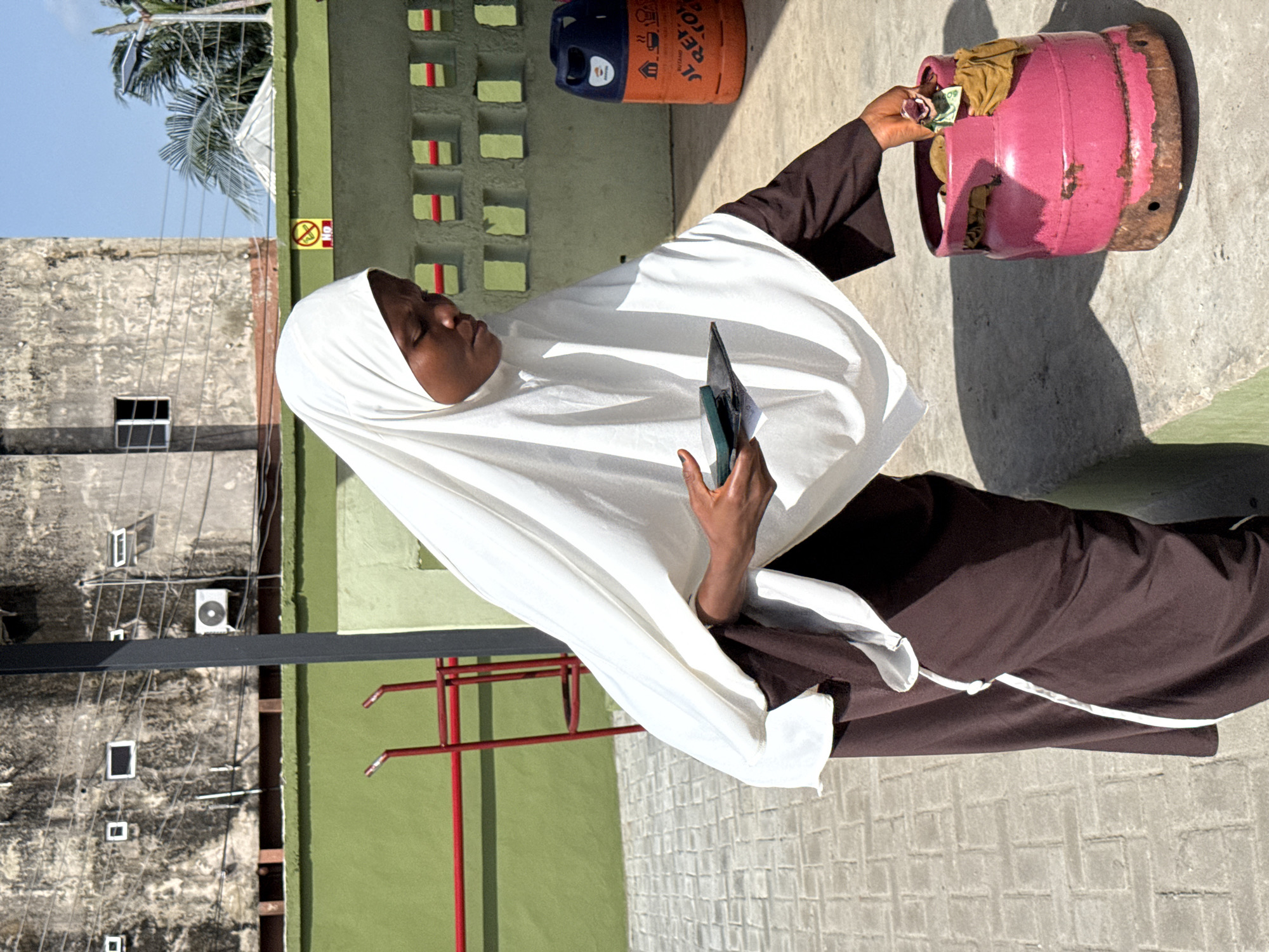 A woman in Ado, Nigeria, wearing a white hijab and brown dress, stands on a concrete surface. She is holding a black wallet in her left hand and a pink gas cylinder in her right hand. The cylinder contains cash and tan-colored fabric. An orange gas cylinder sits in the background, along with a green and yellow wall. The sky is blue, and a building with an air conditioning unit and window is visible.