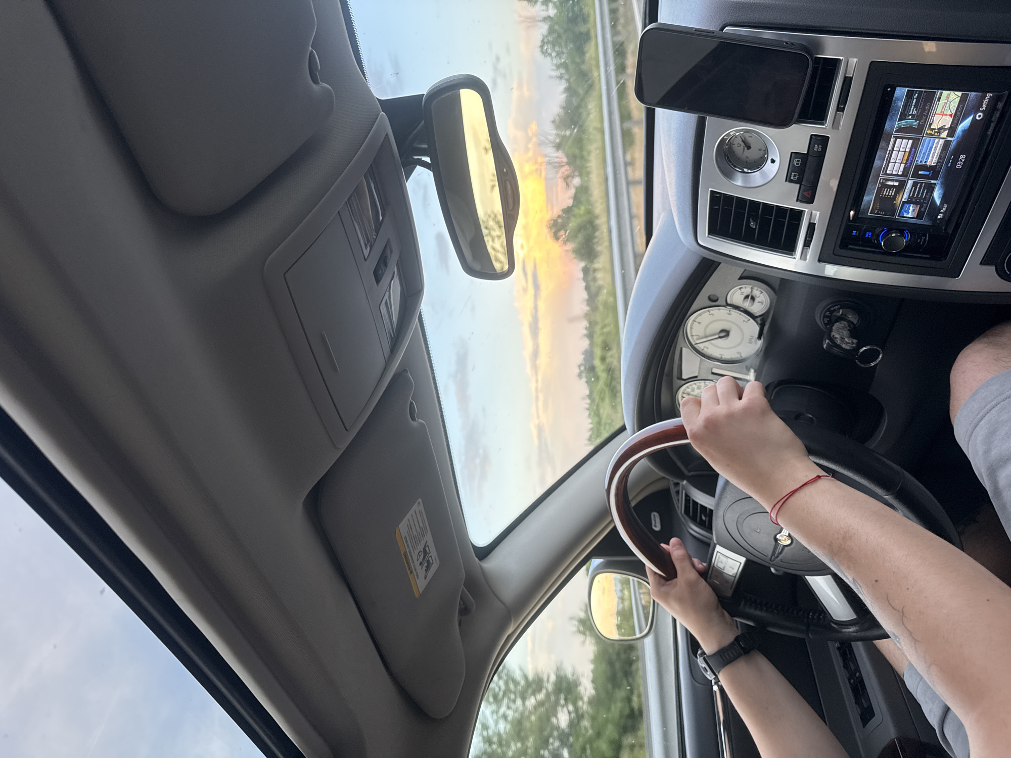 The image offers a first-person perspective from inside a car, likely a Chrysler 300, traveling in Shtarkovo, Bulgaria, during a sunset. The driver's hands, one with a red string bracelet and the other wearing a dark-strapped watch, are seen gripping a wood-grain and black steering wheel. A partial view of a passenger's leg is visible in the lower right.
The car's interior features a gray dashboard with chrome accents, an analog clock, and air vents. An aftermarket car stereo with a digital display shows "Setting," "03:28," and "Rear," indicating various functions. A smartphone is mounted above the central console, and an "ESP OFF" button is visible on the dashboard. Through the windshield, the sky is awash with the warm orange and yellow hues of a sunset, which is also reflected vividly in the rearview mirror. The roadside scenery consists of green trees and foliage, suggesting a drive along a highway or rural road. A sun visor on the passenger side is flipped up, displaying a "WARNING" label with additional, unreadable text. The scene conveys a peaceful journey as dusk settles.