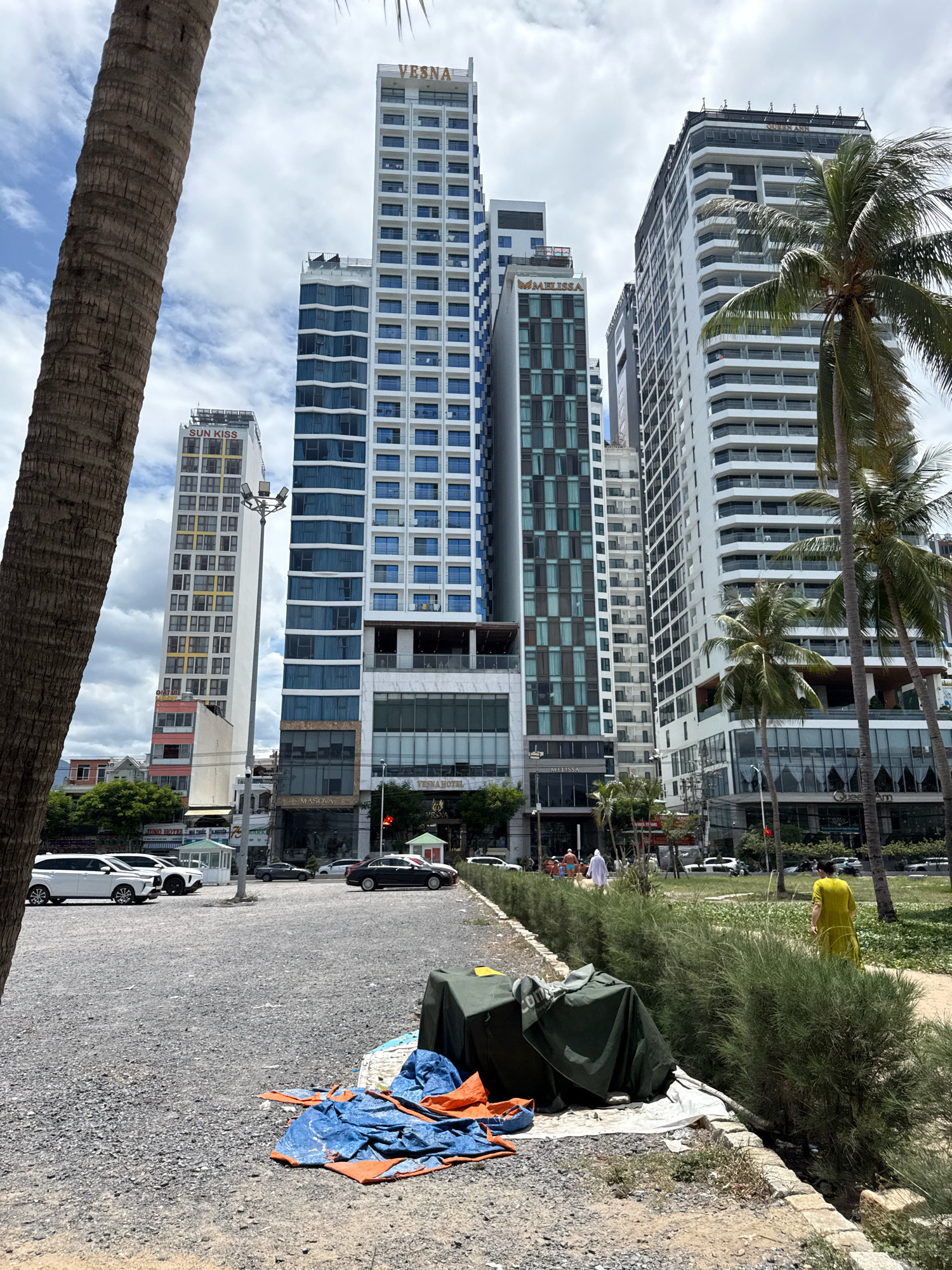 A street scene in Nha Trang, Vietnam, features a foreground of gravel with a few scattered tarps and personal belongings. A line of green bushes separates the gravel area from a pathway and a grassy area, where a woman in a yellow dress is walking away from the camera. In the background, a row of modern high-rise buildings, including the "Vesna Hotel," "Sun Kiss," and "Melissa," line the street. Several cars are parked and driving on the road in front of the buildings. Palm trees are visible throughout the scene. The sky is partly cloudy.