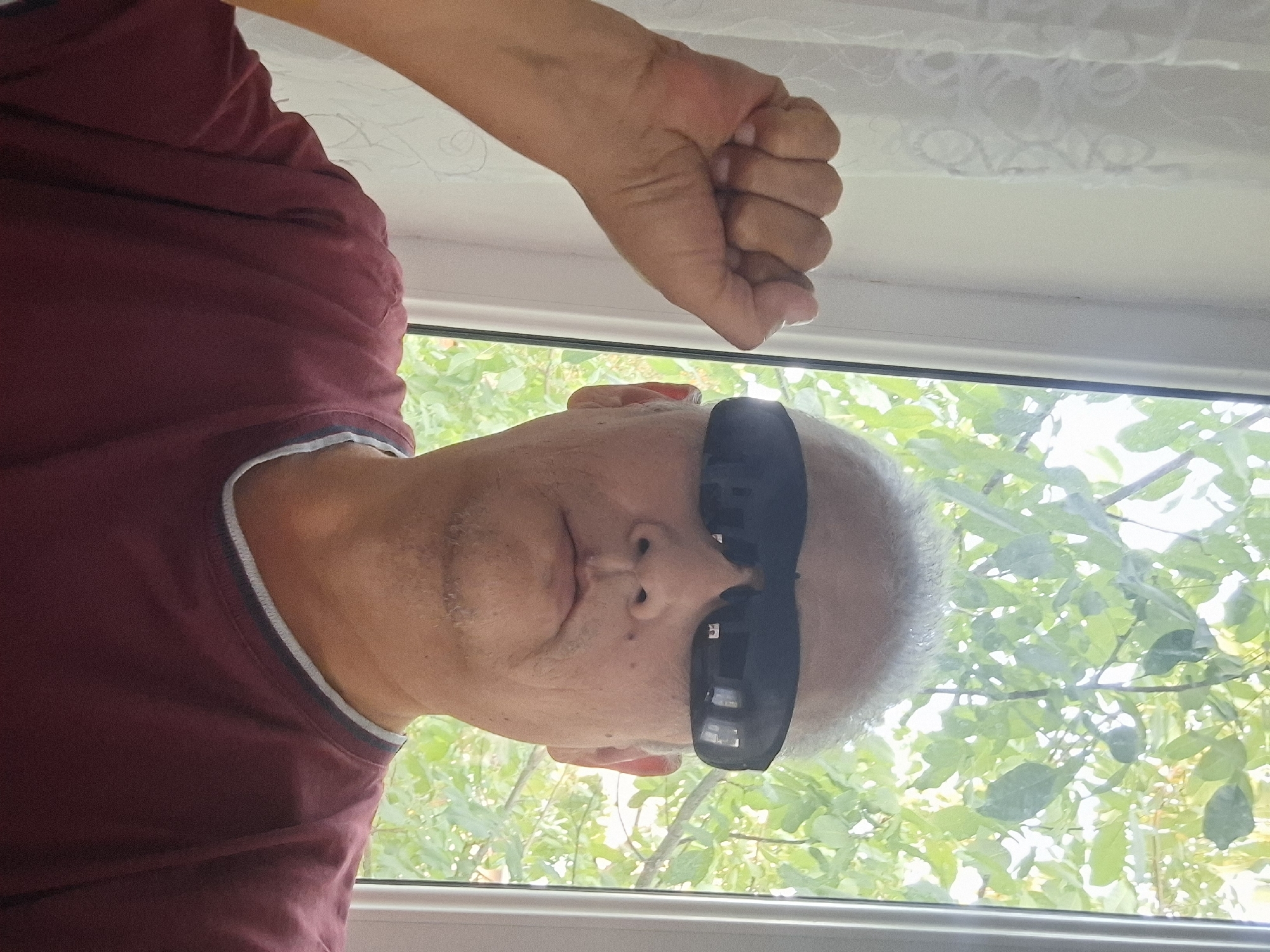 An individual is taking a selfie in front of a window. The person has short gray hair, is wearing dark sunglasses and a maroon t-shirt. Their left arm is raised with a closed fist. Through the window, green foliage is visible, suggesting an outdoor environment. The setting is indoors, possibly a home or apartment, given the curtain on the left side of the frame and the window framing. The image does not provide any specific information about the city of Almaty, Kazakhstan, or any particular urban landmarks.