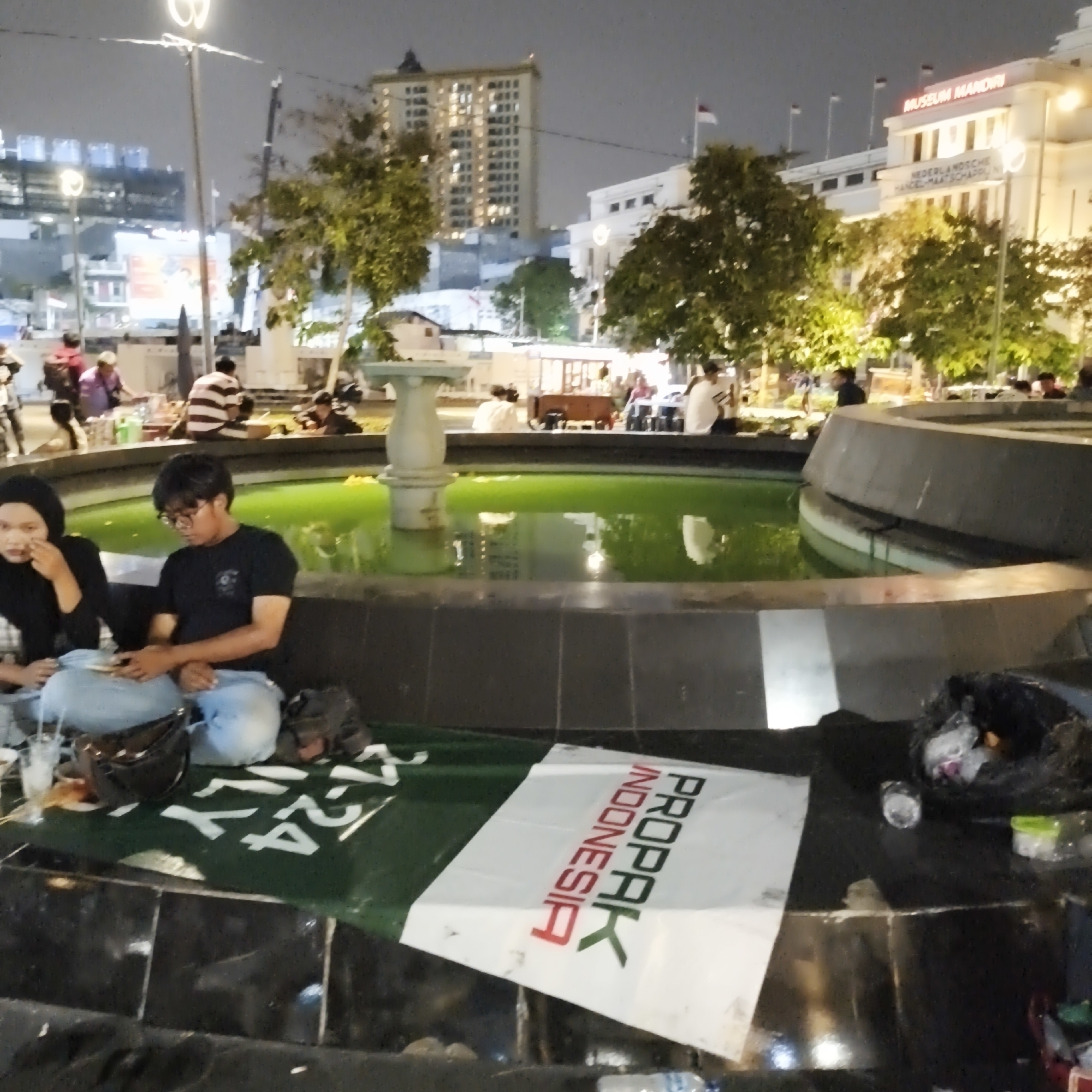 The image shows an outdoor scene at night, likely in an urban area of West Jakarta, Indonesia, near a fountain. In the foreground, two individuals are seated on the ground, with a green banner spread out in front of them. One person, wearing a black hijab and black top, appears to be looking towards their left. The other person, a male wearing glasses and a black t-shirt, is looking down and holding a smartphone. Their legs are covered by blue jeans. To their right, on the ground, is a black garbage bag filled with items and a small plastic container.
The fountain's water is a vivid green color. Behind the fountain, several trees with visible foliage are illuminated. In the mid-ground, there are other people sitting around the fountain and in the background.
The background features several buildings. One building on the left is tall and illuminated, with numerous windows. To the right of this, a white building with a sign that reads "MUSEUM MANDIRI" is visible, along with flags flying on poles. The overall atmosphere suggests a public gathering or event taking place at night.