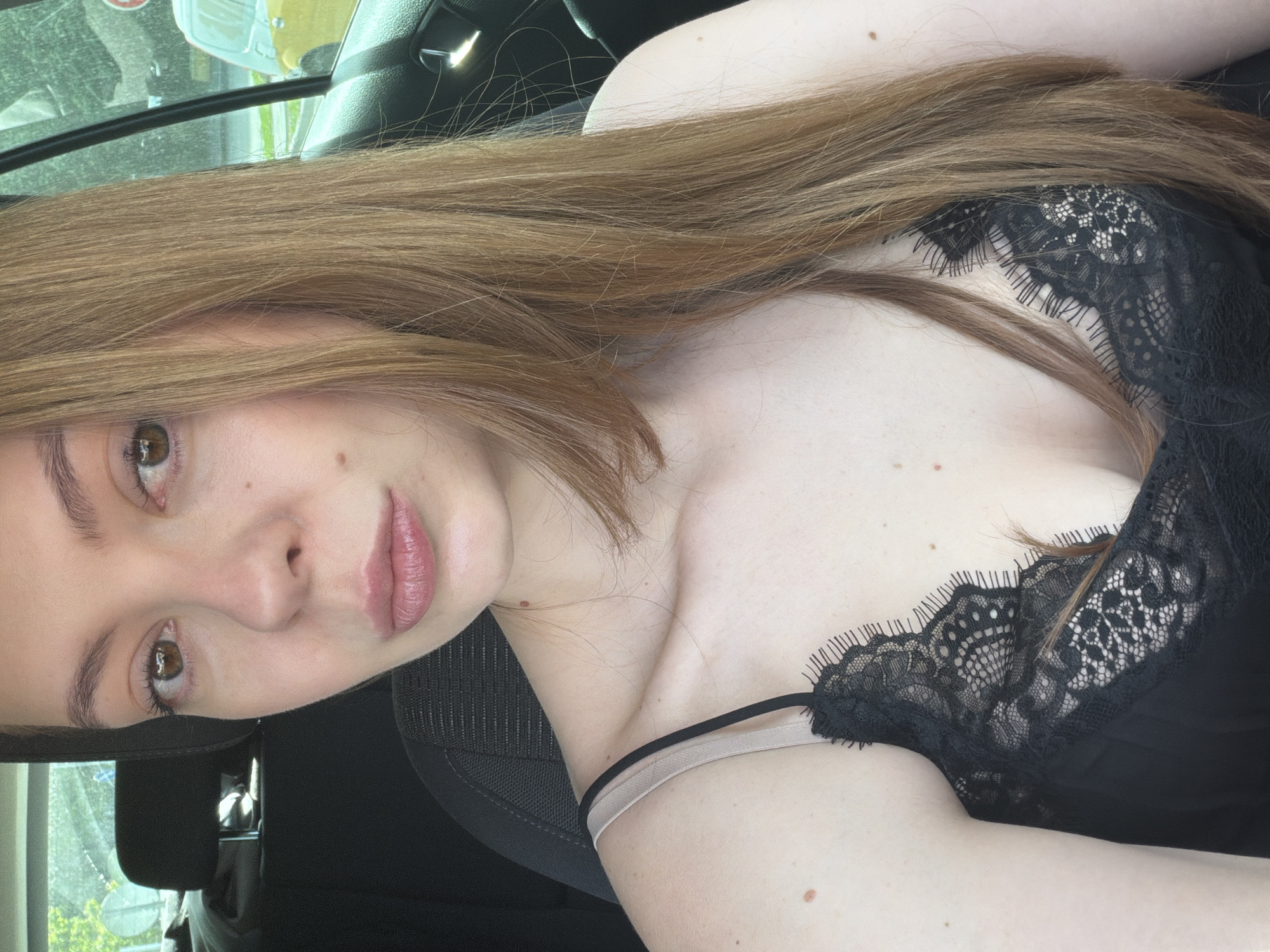 A young woman with light brown hair and fair skin is pictured in what appears to be the interior of a car. She is wearing a black lace top with thin straps. The image is oriented vertically, and she is positioned slightly off-center. The background includes the car's interior and the blurred view outside the window, possibly showing foliage. There's no indication of the location being in Khimki, Russia, based on the image itself.