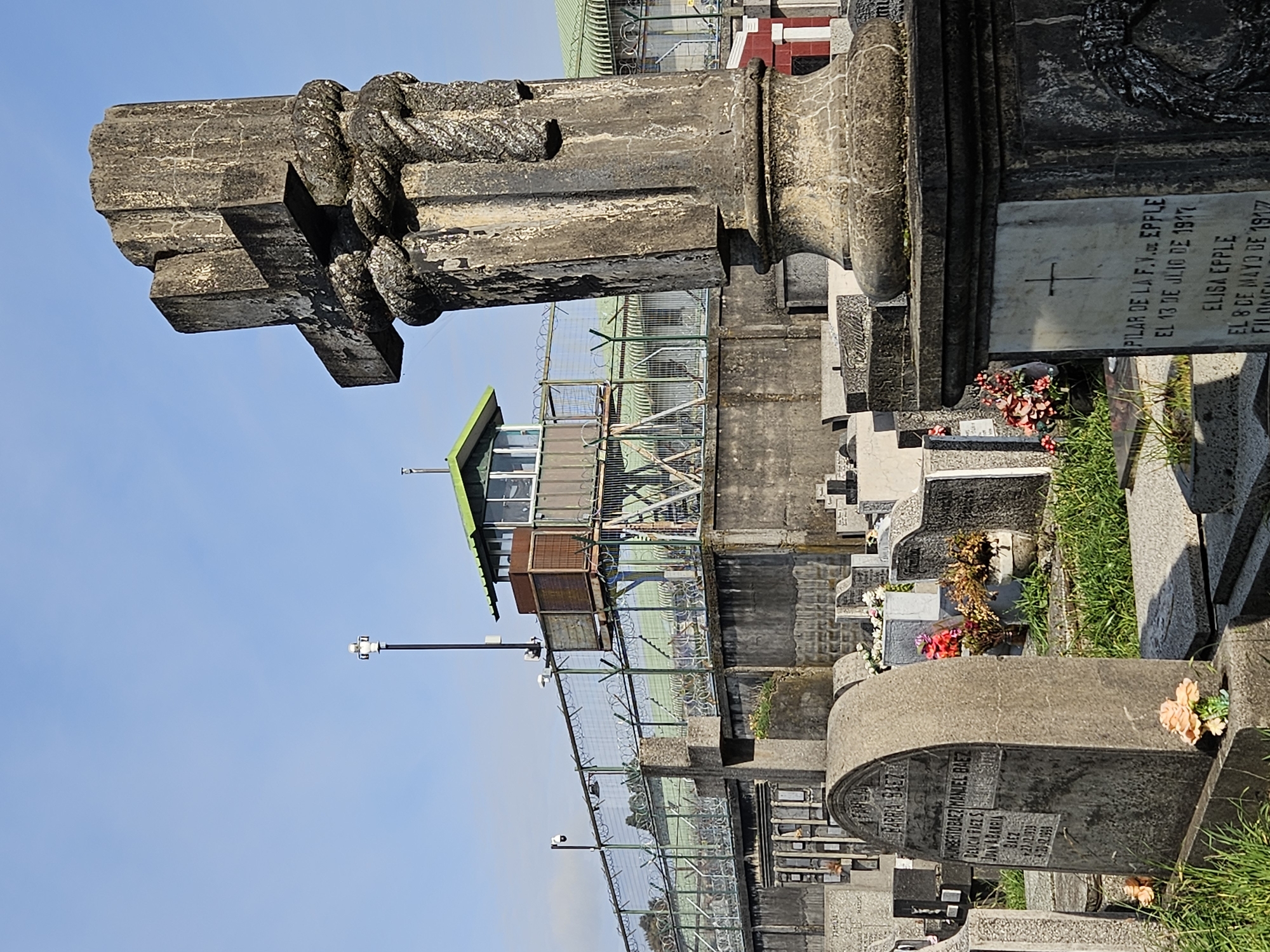 The image depicts a section of a cemetery, possibly in Osorno, Chile, considering the Spanish inscriptions on the gravestones. The foreground features weathered tombstones, some adorned with flowers. Inscriptions on the stones indicate names (e.g., "BARRA BAEZ," "PILAR DE LA F.V.DE EPPLE") and dates of death (e.g., "EL 13 DE JILIO DE 1917," "EL 8 DE MAYO DE 1917"). A large concrete cross, weathered and appearing aged, stands prominently in the frame. A watchtower can be seen behind the tombstones, indicating that this cemetery may be under some form of surveillance or security. A fence with barbed wire runs through the middle ground, likely demarcating different sections of the cemetery or providing security. The sky is clear and blue. Street lamps are visible along the perimeter fence.