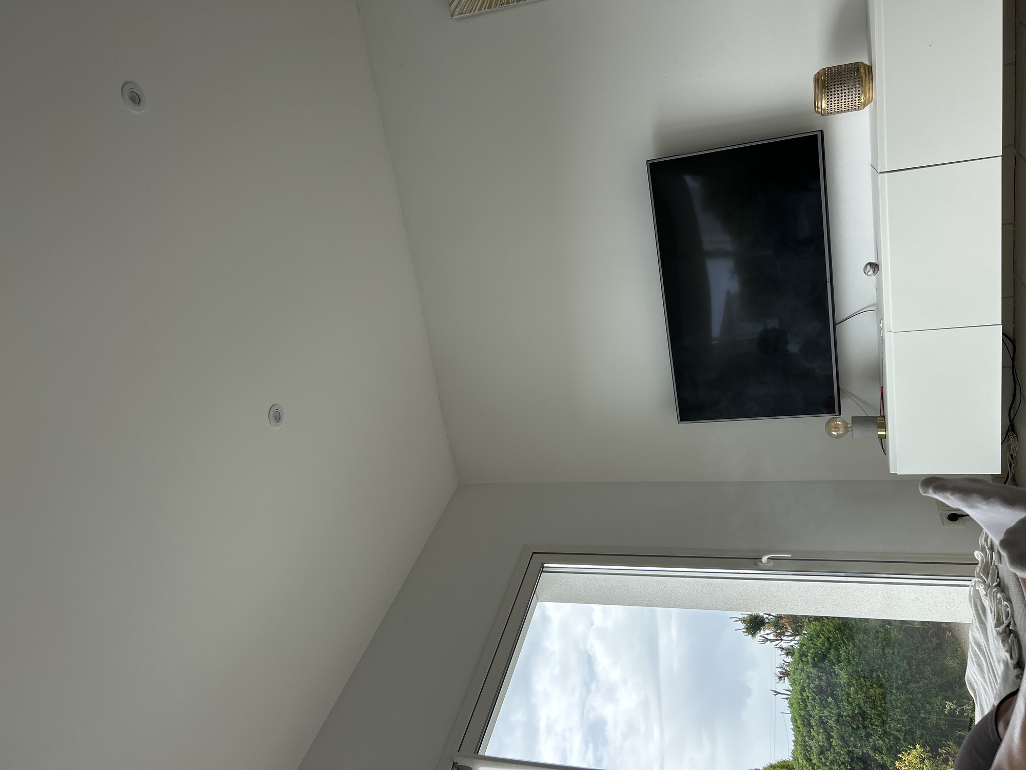 A low-angle interior shot shows a corner of a room. The white ceiling has two recessed lights. A flat-screen TV is mounted on the wall, next to a white storage unit. Below, a window reveals a view of trees and a cloudy sky. Part of a bed with bedding is visible in the lower right corner. The location may be in Veauchette, France, but this cannot be confirmed from the image alone. There are no people visible, only the presence of a foot under the bedding. The primary objects are the TV, storage unit, window, and bed.