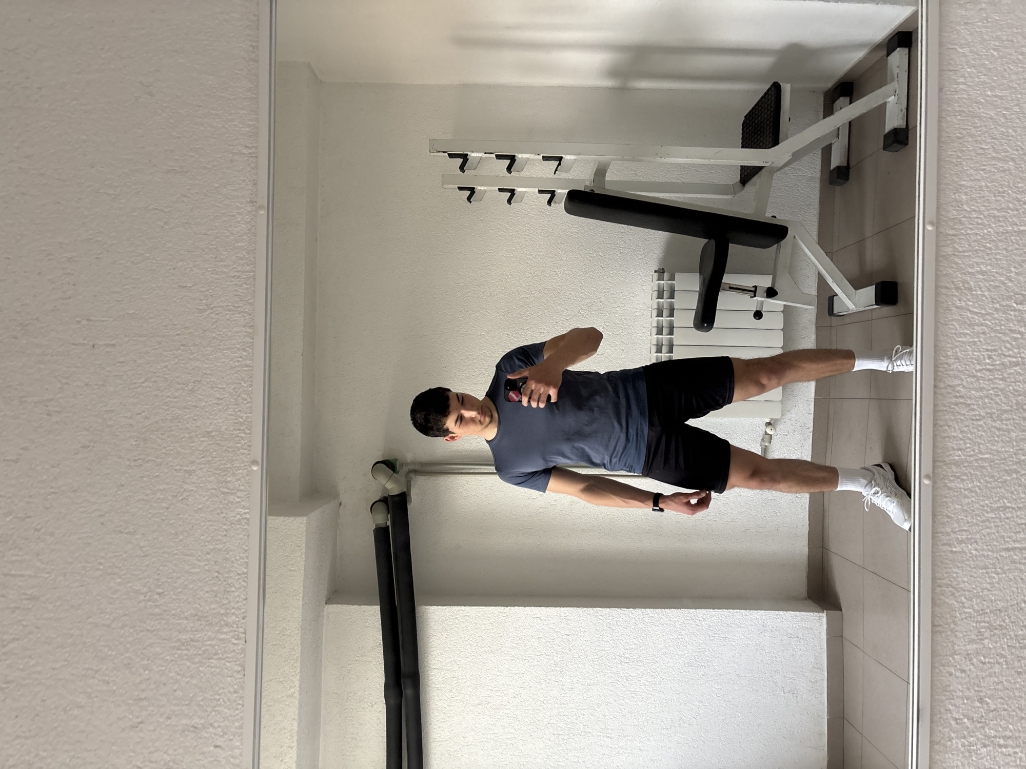The image captures a man taking a mirror selfie in an indoor setting, likely a home gym or a dedicated exercise space in Zlatograd, Bulgaria. The man, positioned centrally in the reflection, is giving a thumbs-up gesture and holding a smartphone, with a slight smile on his face. He is dressed in a dark blue short-sleeved t-shirt, black shorts, white socks, and white athletic shoes, with a dark watch visible on his left wrist.
The background, also seen in the mirror, features various pieces of exercise equipment. To the man's right, there is a white weight bench with a black padded seat and backrest, alongside a white rack equipped with multiple hooks or holders, possibly for barbells or weights. A white radiator is visible behind the weight bench. The walls are textured and light-colored, appearing off-white or light grey. The floor is tiled in a light neutral tone. In the corner to the man's left, two dark, insulated pipes run vertically along the wall. The overall scene suggests a clean, functional space dedicated to fitness. No discernible text is visible within the image or its crops.
