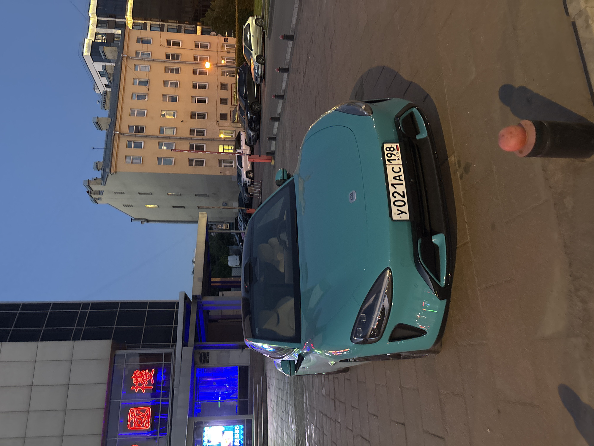 A teal-colored electric car, possibly a NIO ET7, is parked on a paved area in front of a building with neon signs in Chinese characters. The car has a black roof and its headlights are on. The license plate reads "y021AC 198 RUS". In the background, there are several buildings, including a light yellow, multi-story apartment building with many windows and a modern glass-fronted building. The sky is a dusky blue, suggesting it is either early morning or late evening. The scene appears to be in a urban environment, likely a city street or parking lot. The overall impression is a contemporary urban setting with a focus on the electric vehicle.