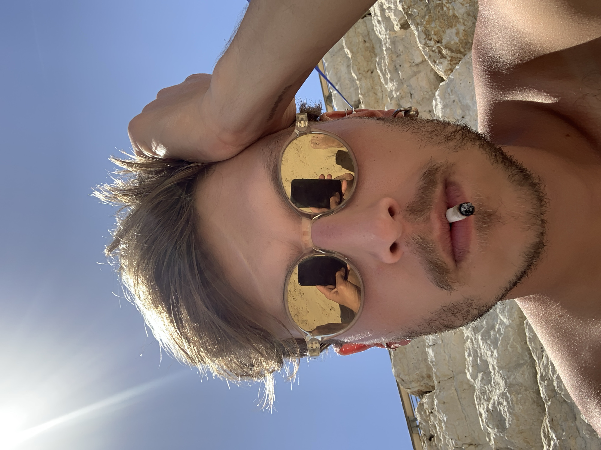 A man with a light complexion, brown hair, and a light beard and mustache is seen from a low angle, with a bright blue sky partially visible in the background. He is wearing reflective sunglasses, and a cigarette is held between his lips. His left hand is raised and appears to rest on his head. The sunglasses reflect the person holding the phone to take the picture. Part of a textured, light-colored structure, possibly a stone wall, is visible on the right. The location is Tel-Aviv, Israel.