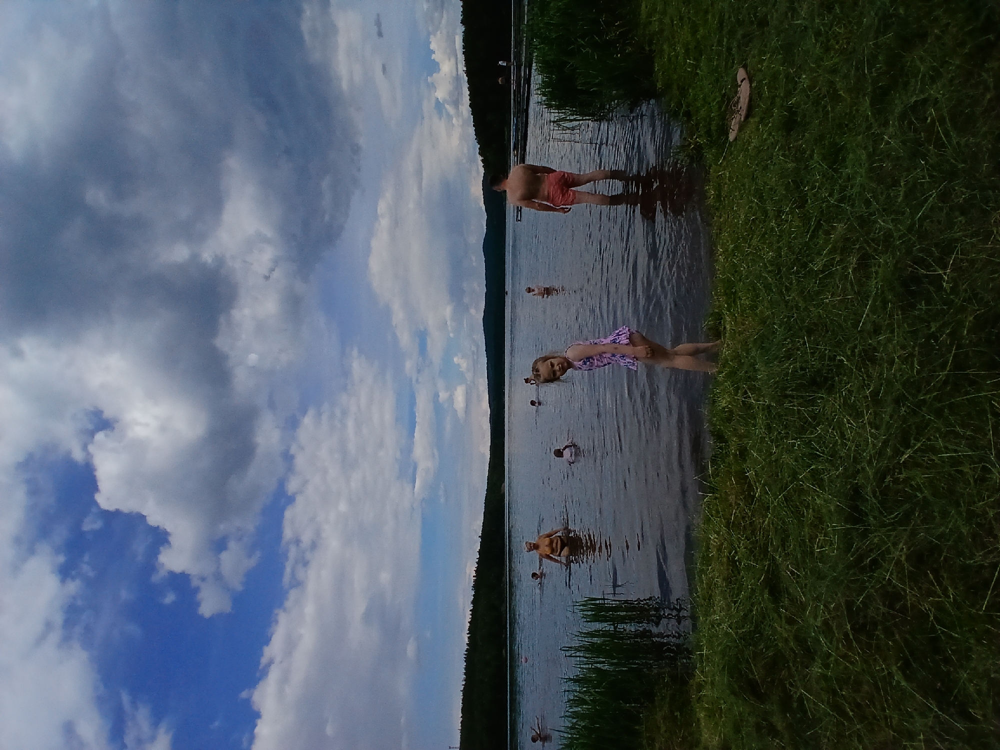 A wide shot captures a cloudy, overcast day at a lake, possibly in Revda, Russia, where several people are swimming and wading. In the foreground, lush green grass lines the bank, with a pair of sandals visible on the right. The water is calm and dark, reflecting the dramatic sky. Several individuals are scattered across the lake. A man in coral-colored shorts stands in the water on the right, facing away from the camera. To his left, a young girl in a patterned swimsuit faces the viewer, her gaze directed slightly away. Further out in the water, various people are swimming, some with their heads visible above the surface, and others appearing to be farther from shore. The background features a dense line of dark green trees along the horizon, with hills faintly visible beyond. The sky is dominated by large, grey clouds with patches of blue peeking through, suggesting a dynamic weather pattern.