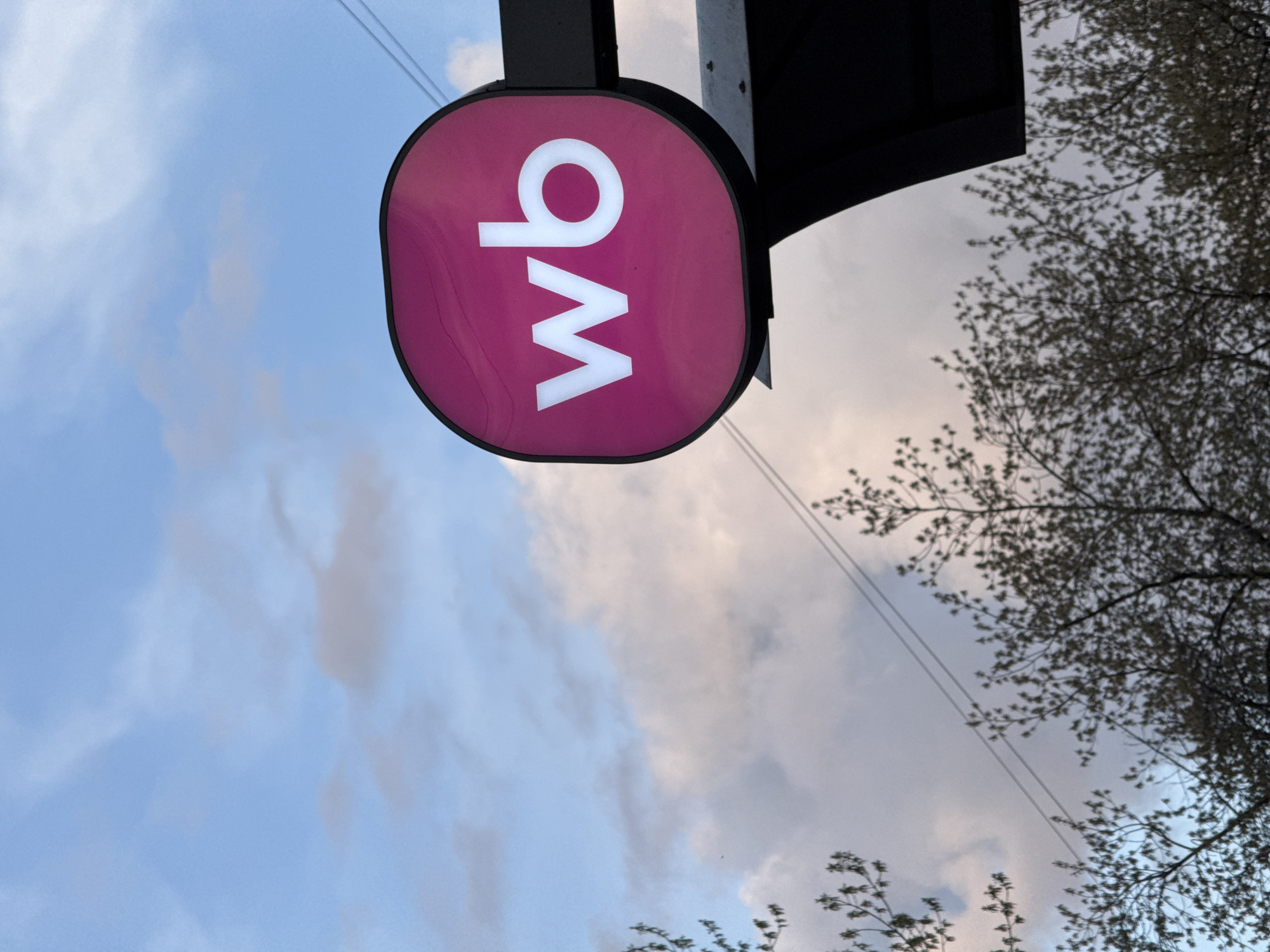 The image features a pink sign with the letters "wb" in white, attached to a dark metal structure, likely a building. The sign is situated against a backdrop of a cloudy sky and bare tree branches. This sign is for Wildberries, the Russian e-commerce company.
Without additional context or identifiers on the image itself, pinpointing the exact location within Krasnoyarsk is not possible based on visual elements alone.