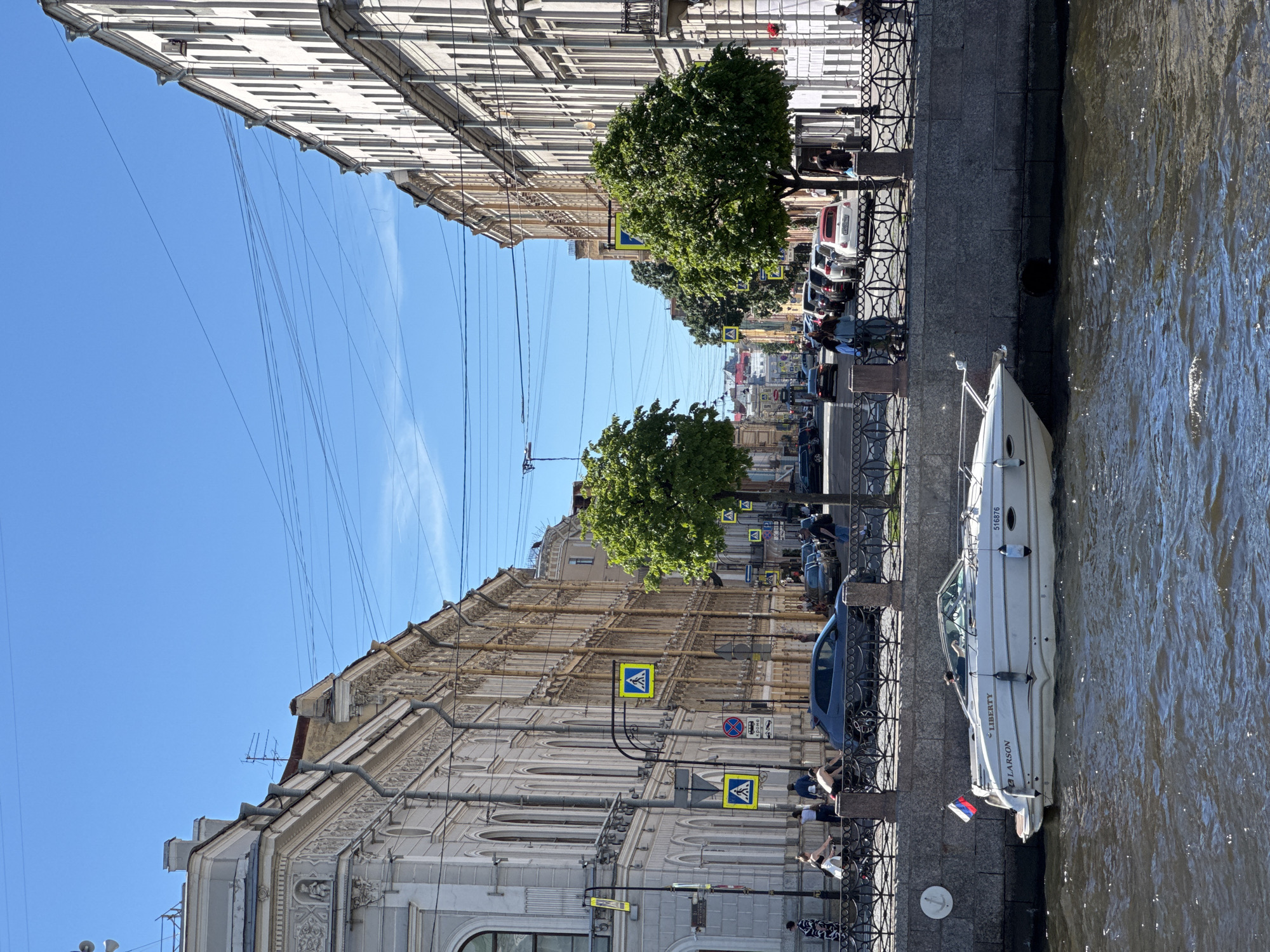 This image captures a bright, sunny daytime scene along the Fontanka River embankment in Saint Petersburg, Russia. In the immediate foreground, the dark, rippling water of the Fontanka River occupies the lower third of the frame. A white Larson Liberty motorboat, bearing the hull number "516876" and flying a Russian flag from its stern, is moored alongside a grey stone embankment on the right. One person is visible in the boat's cockpit. An circular anchor relief is affixed to the stone embankment wall.
Above the embankment, a black ornate metal fence separates the waterway from a paved street. Along this street, multiple cars are parked and moving, including a prominent white Fiat 500 convertible with a red fabric roof. Several pedestrians are scattered along the sidewalk, some walking, others standing, and two individuals appear to be on bicycles on the left side of the street. Two mature green deciduous trees with full foliage are positioned along the sidewalk, contributing to the midground. Street furniture includes black lamp posts, some of which display yellow and blue pedestrian crossing signs, and a red and blue circular "no stopping" sign with Russian text "КРОМЕ" (KROME) indicating exceptions.
Both sides of the street are lined with multi-story classical-style buildings featuring intricate architectural details, such as decorative moldings, cornices, and numerous arched and rectangular windows. Some buildings show drainpipes and rooftop antennas against a clear, bright blue sky, which dominates the upper portion of the image. The street recedes into the background, revealing more of the city's architecture under bright sunlight, indicating clear weather.