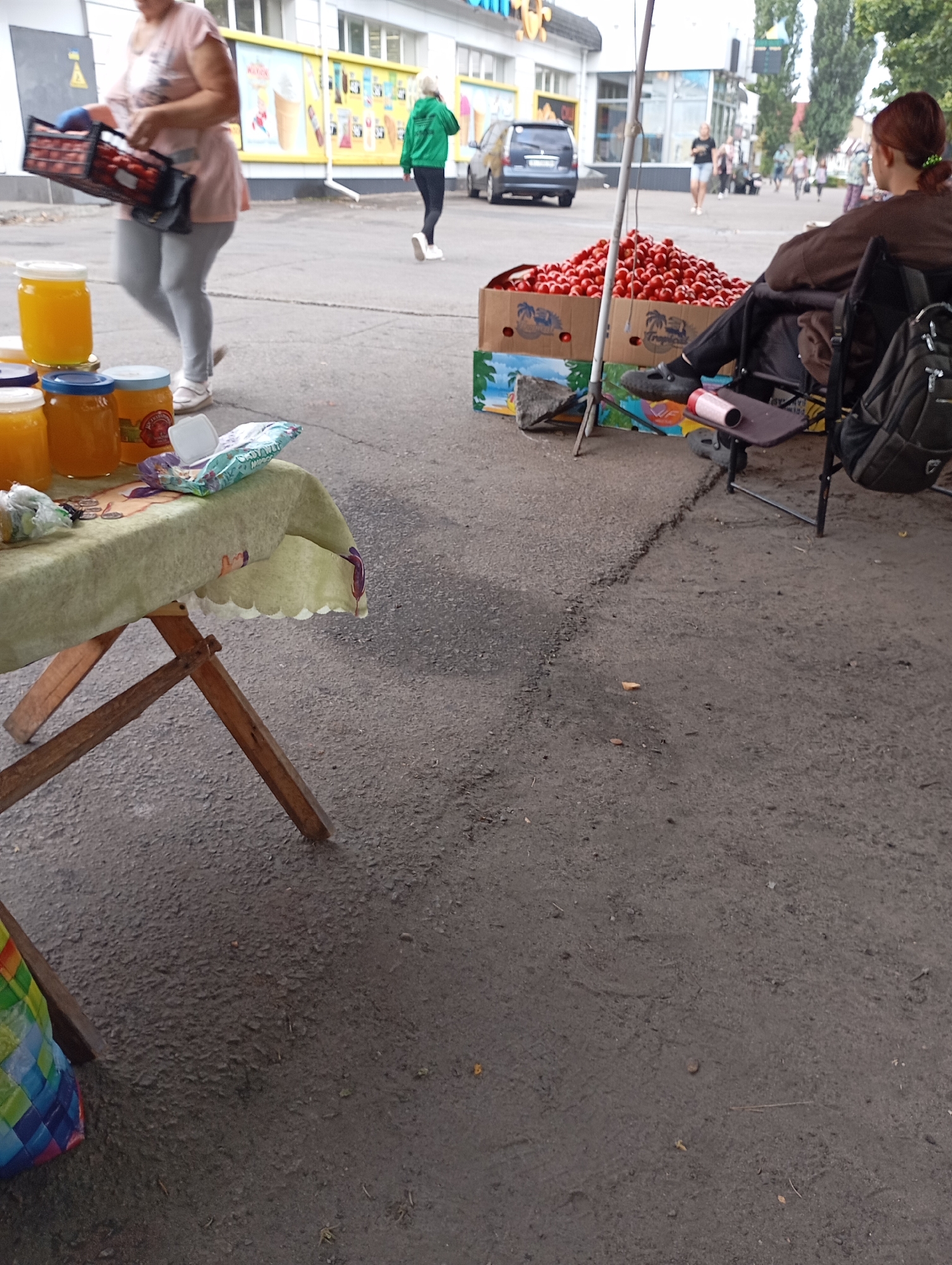 The image captures a street scene in Horishni Plavni, Ukraine, likely a market or roadside stall. In the foreground, a wooden folding table covered with a light green tablecloth displays several jars of honey and a colorful woven bag. To the right, a person sits in a folding chair next to a large cardboard box overflowing with red tomatoes. Another individual is seen in the background carrying a crate of tomatoes. A person in a green hoodie walks away from the camera towards a building with advertisements on its facade. A dark colored car is parked on the street. The ground is paved with asphalt, and trees are visible in the distance. The overall atmosphere suggests a commercial exchange taking place in an outdoor urban setting.