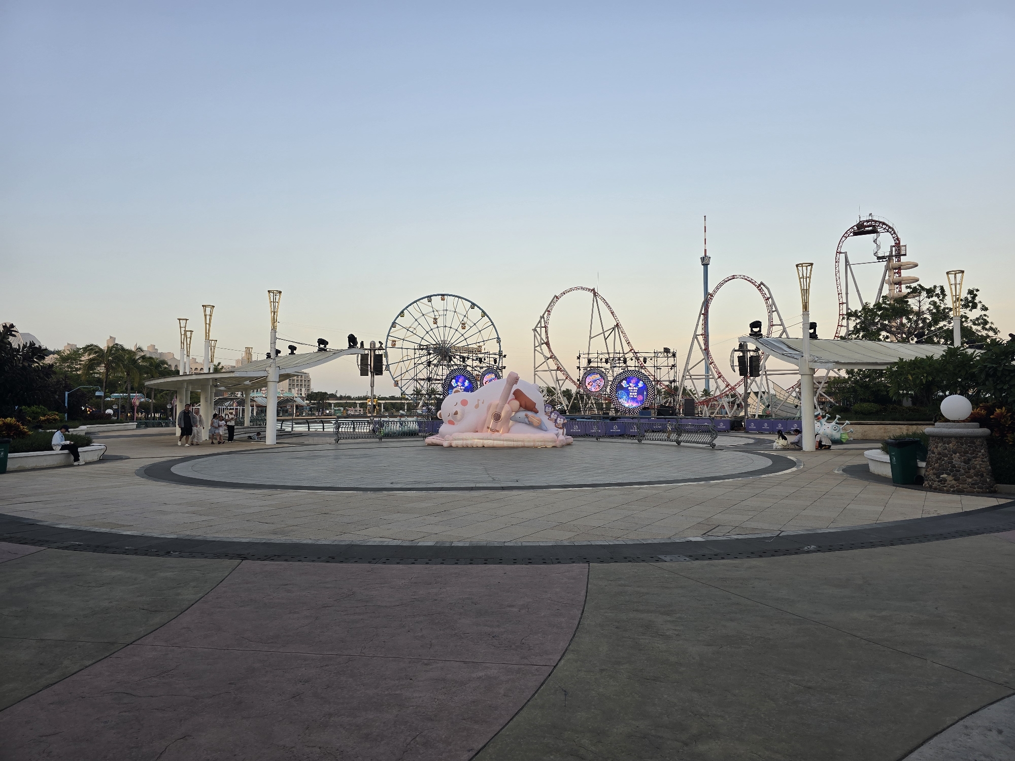 The image depicts an outdoor plaza in front of amusement park attractions at dusk. In the foreground, a circular paved area features distinct zones of gray and purple concrete, bordered by dark gray concrete strips with a textured pattern. A large, pale pink inflatable sculpture of a sleeping bear holding a guitar is centrally located in the plaza, surrounded by decorative lighting.
In the mid-ground, a white shaded structure with tall, ornate pillars provides shelter. Several people are visible walking and sitting under this structure and along a low wall. Beyond this, the backdrop is dominated by amusement park rides, including a Ferris wheel on the left and several roller coasters with red tracks and white supports. Illuminated circular signs are visible on the roller coaster structures.
The background shows more of the amusement park structures and some trees and buildings. The sky is a clear, pale blue, indicating early evening or late afternoon. The overall scene suggests a recreational area, likely a theme park or amusement park, situated in the city of Li'an, China.