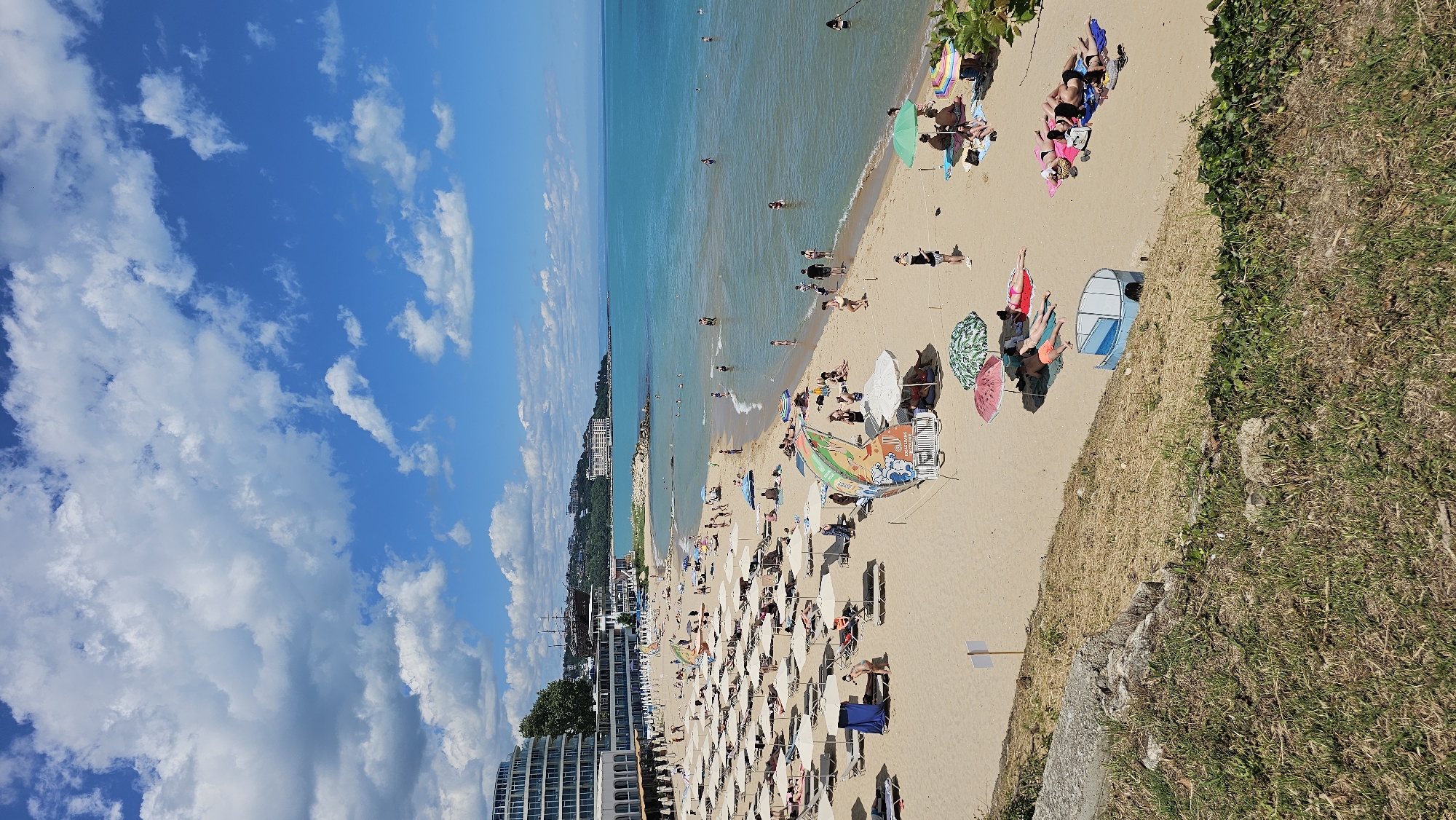 The image depicts a bustling beach scene on a sunny day. The location is likely the central beach of Varna, Bulgaria, known for its long sandy stretch and clear blue waters. In the foreground, a grassy embankment with some dry vegetation overlooks the beach. The beach itself is densely populated with numerous beach umbrellas and loungers, indicating a high volume of visitors. People are scattered across the sand, some sunbathing on towels, others sitting under umbrellas, and some are in the shallow waters of the sea. The sea is a vibrant turquoise, with gentle waves lapping the shore. In the background, a large hotel complex with a distinctive curved facade dominates the coastline, typical of Varna's resort architecture. Further along the coast, more buildings and a distant hill can be seen. The sky is bright blue with scattered white clouds, contributing to the overall idyllic summer atmosphere. There are various objects visible on the beach, including beach chairs, umbrellas of different colors and patterns, towels, bags, and what appears to be a small child's play structure. The presence of many people engaging in leisure activities suggests a popular tourist destination during peak season.