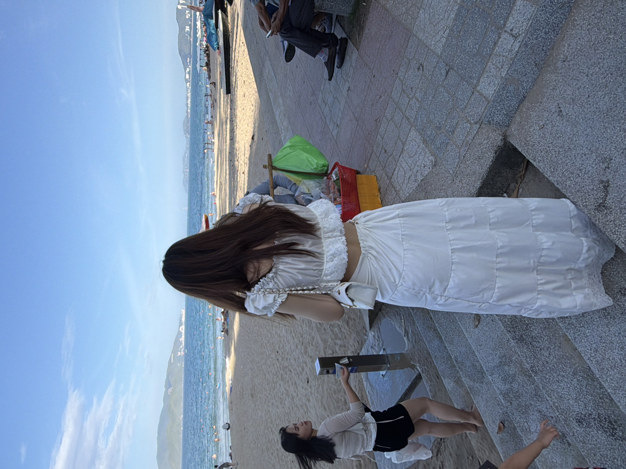 The image depicts a beach scene in Nha Trang, Vietnam, likely at the popular Nha Trang Beach or a nearby resort area. The focus is on a woman in the foreground, seen from behind, wearing a white, two-piece outfit consisting of a cropped ruffled top and a long, tiered skirt. She is standing on a set of stone steps leading down towards the beach. Her dark hair is long and flows down her back.
To her left and slightly below, another woman is walking up the steps, also from behind, wearing a light-colored, long-sleeved top and black shorts. She is holding a white plastic bag in her left hand and appears to be interacting with a metal dispenser on a pole.
In the background, the sandy beach is dotted with people sunbathing and swimming in the blue ocean. The water is calm with buoys indicating swimming areas. Further in the distance, hills and a coastline with buildings are visible under a bright blue sky with some scattered clouds. On the paved walkway to the right of the steps, a red basket filled with items and a green plastic bag are visible. Two people are seated on a bench to the far right, one of whom is looking at a mobile phone. The overall impression is a sunny day at a bustling Vietnamese beach.