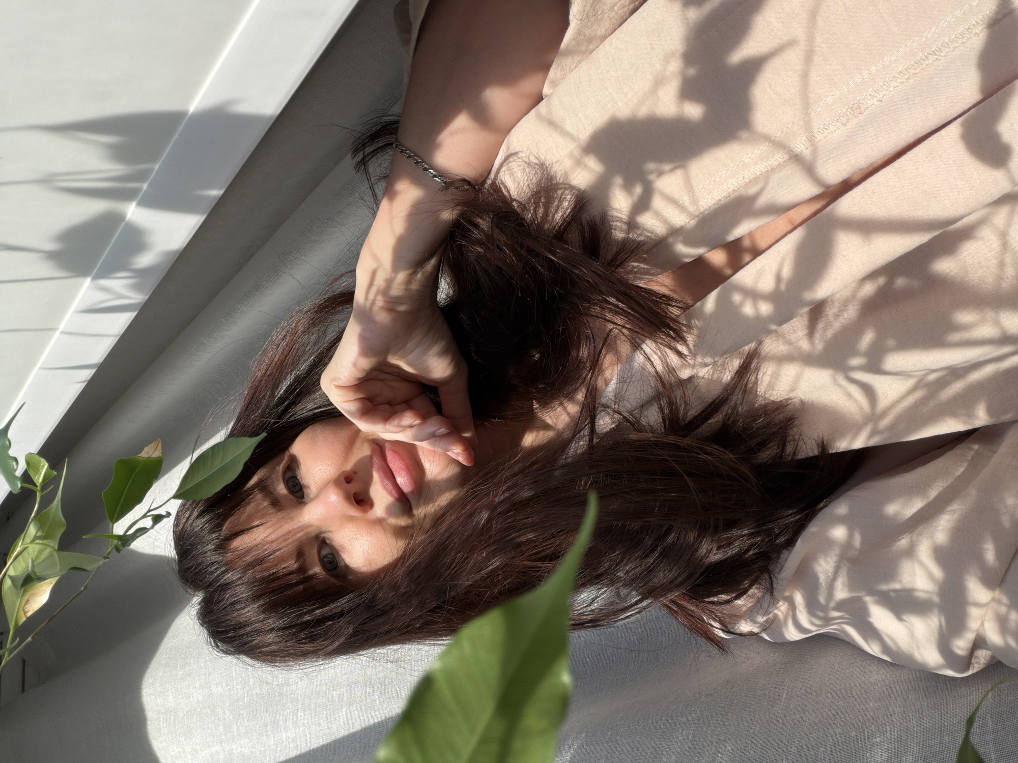 A woman with dark brown hair and green eyes is lying down on a light-colored surface, possibly a bed or a sofa. She is wearing a light beige garment. Her right hand is raised near her face, and she has a silver bracelet on her wrist. There are shadows of plant leaves on the wall and her clothing. A green leafy plant is visible in the lower left corner and foreground of the image. The lighting suggests it is daytime. The image does not provide enough information to determine if it was taken in the city of Perm, Russia.