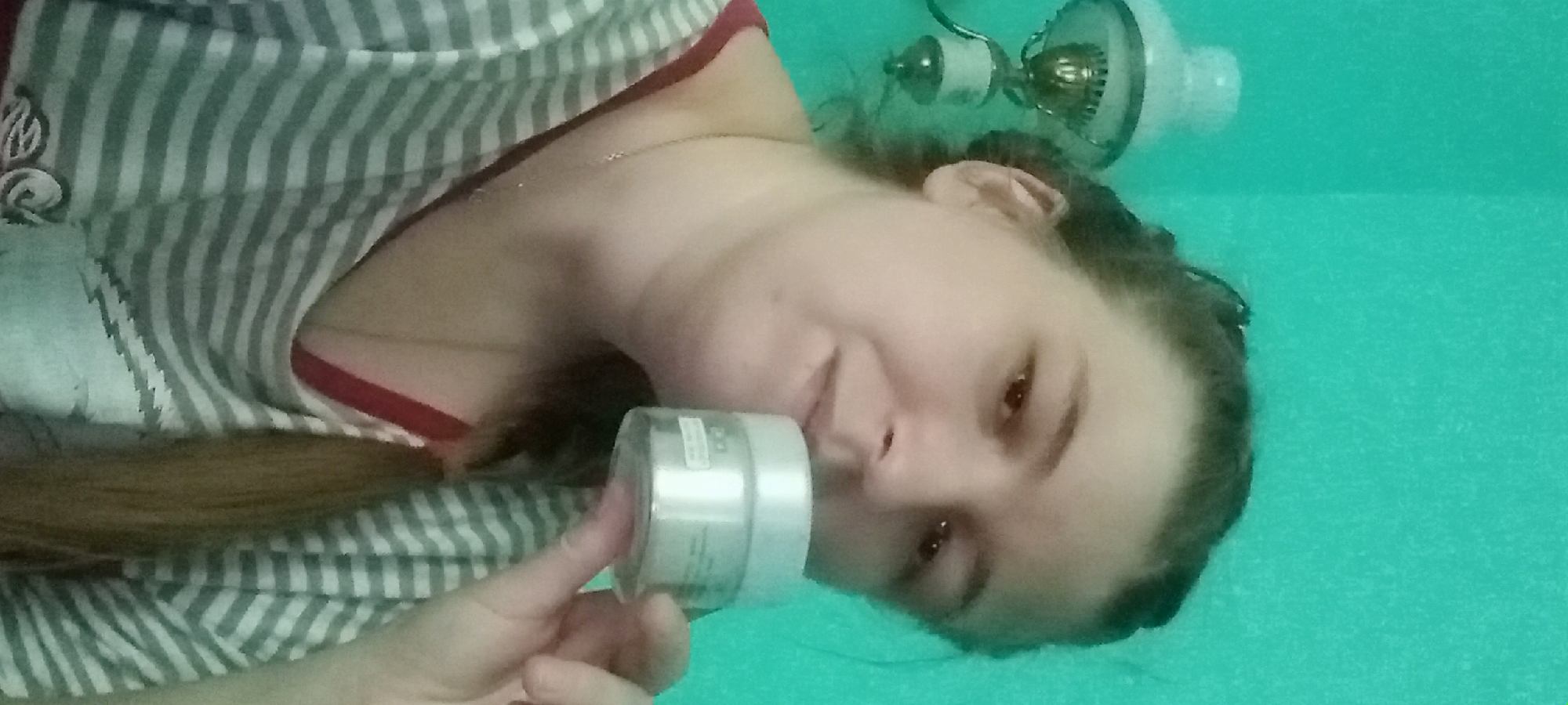 A woman, with her hair pulled back in a braid, holds a small, white jar of skincare product in her right hand, close to her face. She is looking towards the viewer, with a slight smile. She is wearing a grey and white striped t-shirt with a red trim around the neckline. A thin gold necklace with a small pendant is visible around her neck. In the background, to the left of the woman, is a vintage-style oil lamp mounted on the wall. The wall itself is painted a bright, turquoise color, consistent with the typical interior paint choices found in residential buildings in the city of Kitskany, Moldova.