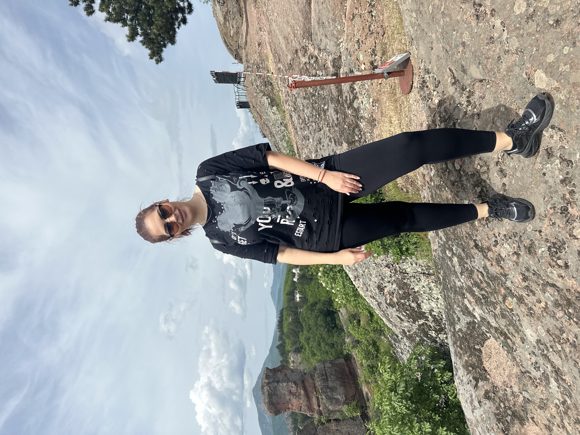 A woman stands on a rocky surface overlooking a landscape in Belogradchik, Bulgaria. She is wearing a black t-shirt, black leggings, and black sneakers. She also has sunglasses. The landscape features rock formations, green trees, and a cloudy sky. A small metal structure is visible on the rocks behind the woman.