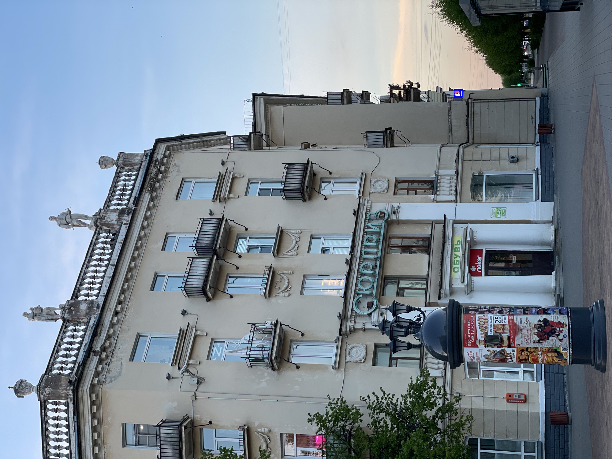 The image shows a multi-story building in Zheleznogorsk, Russia, with a beige facade and decorative elements including statues along the roofline and small balconies. The building houses a shop, identified by a sign reading "ОБУВЬ" (shoes) and the brand "rieker". A sign written in Cyrillic reads "Маки ПЕСНЯ РУССКАЯ- КАК ТЫ ХОРОША" indicating Russian folk culture is celebrated. A large pillar with an attached lamppost displaying posters is in the foreground. The posters appear to advertise musical and cultural events. The sky is visible in the background, suggesting it is either dusk or dawn. The presence of a shop suggests a commercial or populated area within the city.
