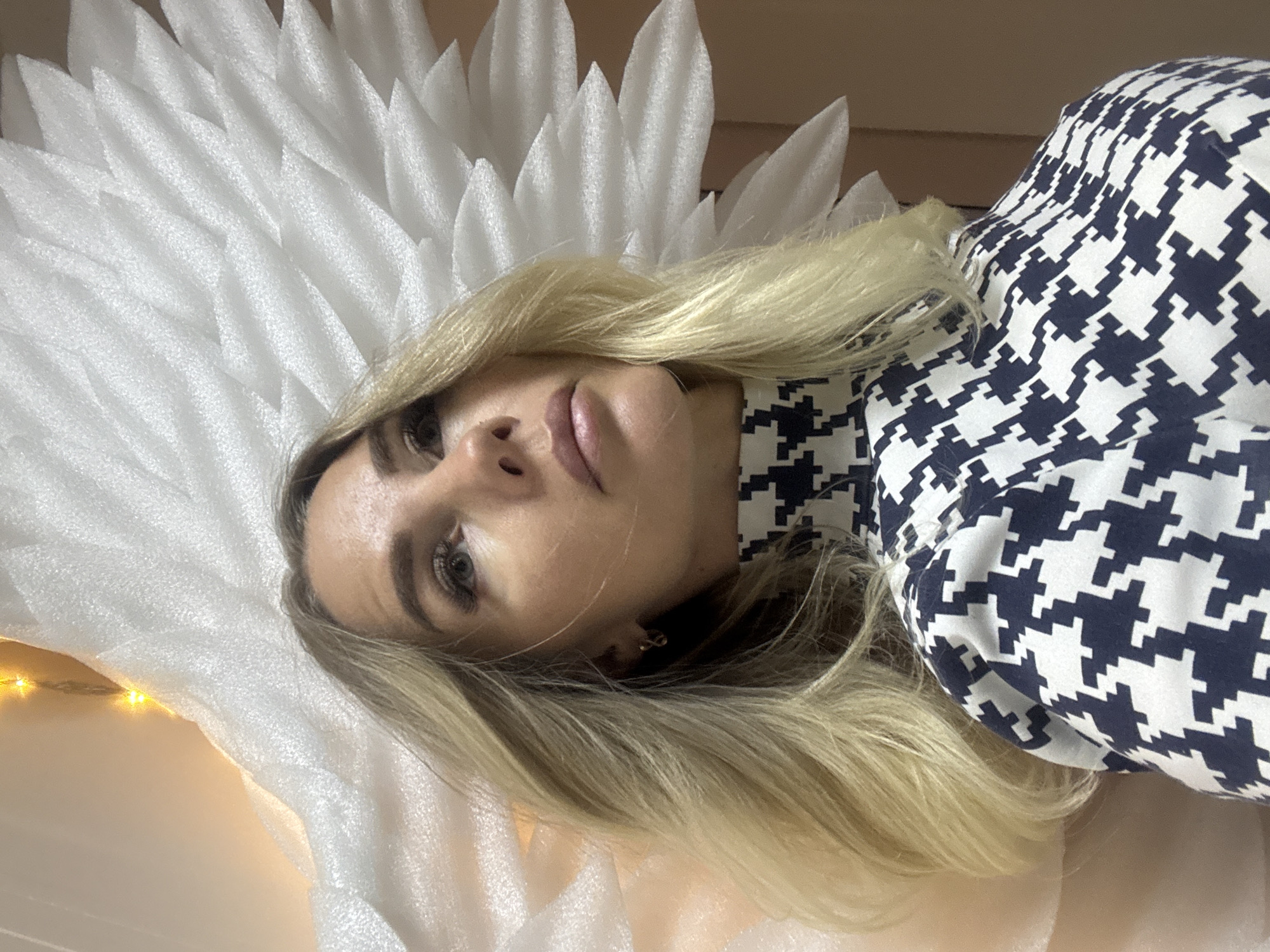 A woman with blond hair and a blue and white houndstooth pattern shirt is lying down. She has fair skin and is wearing earrings. She is lying on a white, textured surface with a feather-like arrangement. In the lower left corner, there are faint yellow lights. The location is not discernible as being in Abakan, Russia.