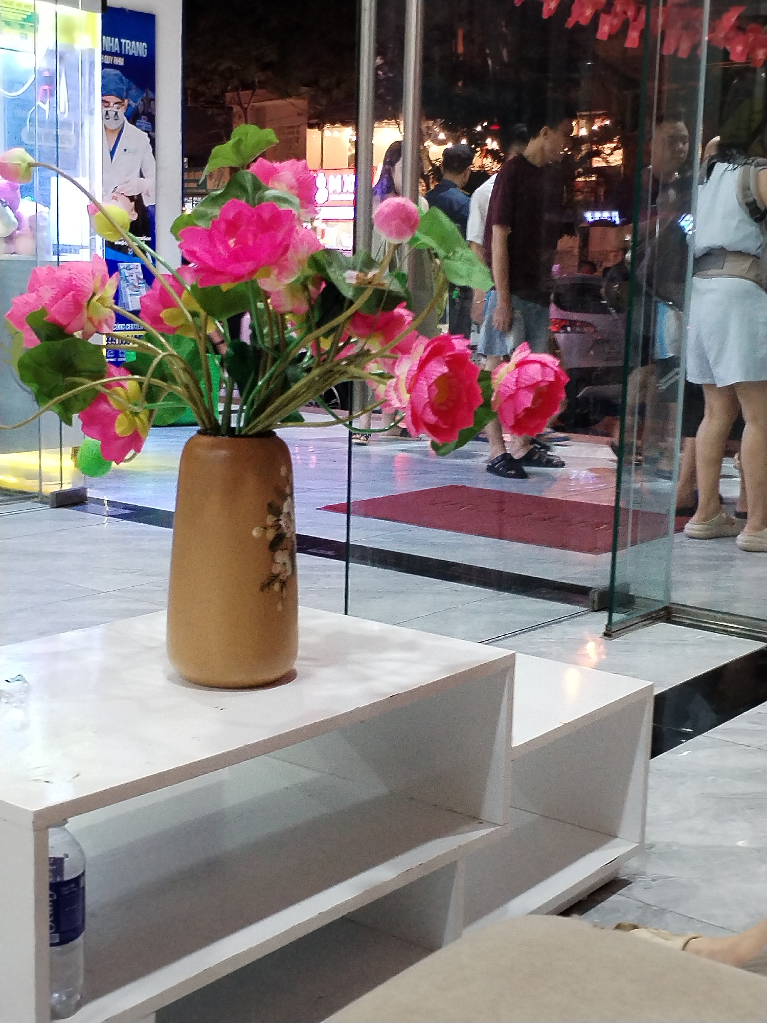 The image captures a night scene, likely an indoor or semi-outdoor public area such as a waiting space or a shop entrance, with a strong sense of artificial lighting.
In the foreground, a multi-tiered white table holds a light brown vase adorned with delicate floral patterns. The vase contains a vibrant bouquet of artificial pink flowers, resembling lotus or peony blooms, with green leaves and some smaller yellow-green buds. A clear plastic water bottle is visible on the lower shelf of the table. In the bottom right corner, a small portion of a person's leg and foot, wearing a light-colored shoe, suggests someone is seated nearby.
Beyond the table, through a clear glass partition, several people are visible in the midground and background. A man in a dark red shirt and blue shorts stands looking to the left. Further back, a woman in a light top and shorts/skirt is partially visible. Other indistinct figures are present, some appearing to walk or stand. The floor visible through the glass is tiled with a red doormat placed on it.
On the left, a blue poster or sign featuring a person in a white lab coat and a blue surgical cap and mask is prominent. The text "NHA TRANG" is clearly visible at the top of this poster, with "QUY NHI" partially visible below it. Further text on the poster is obscured or unreadable. In the background, beyond the people and glass, an urban night environment is suggested by distant lights and dark structures. Red decorative elements are reflected in the glass overhead. The scene suggests a warm climate, given the light clothing worn by the people.