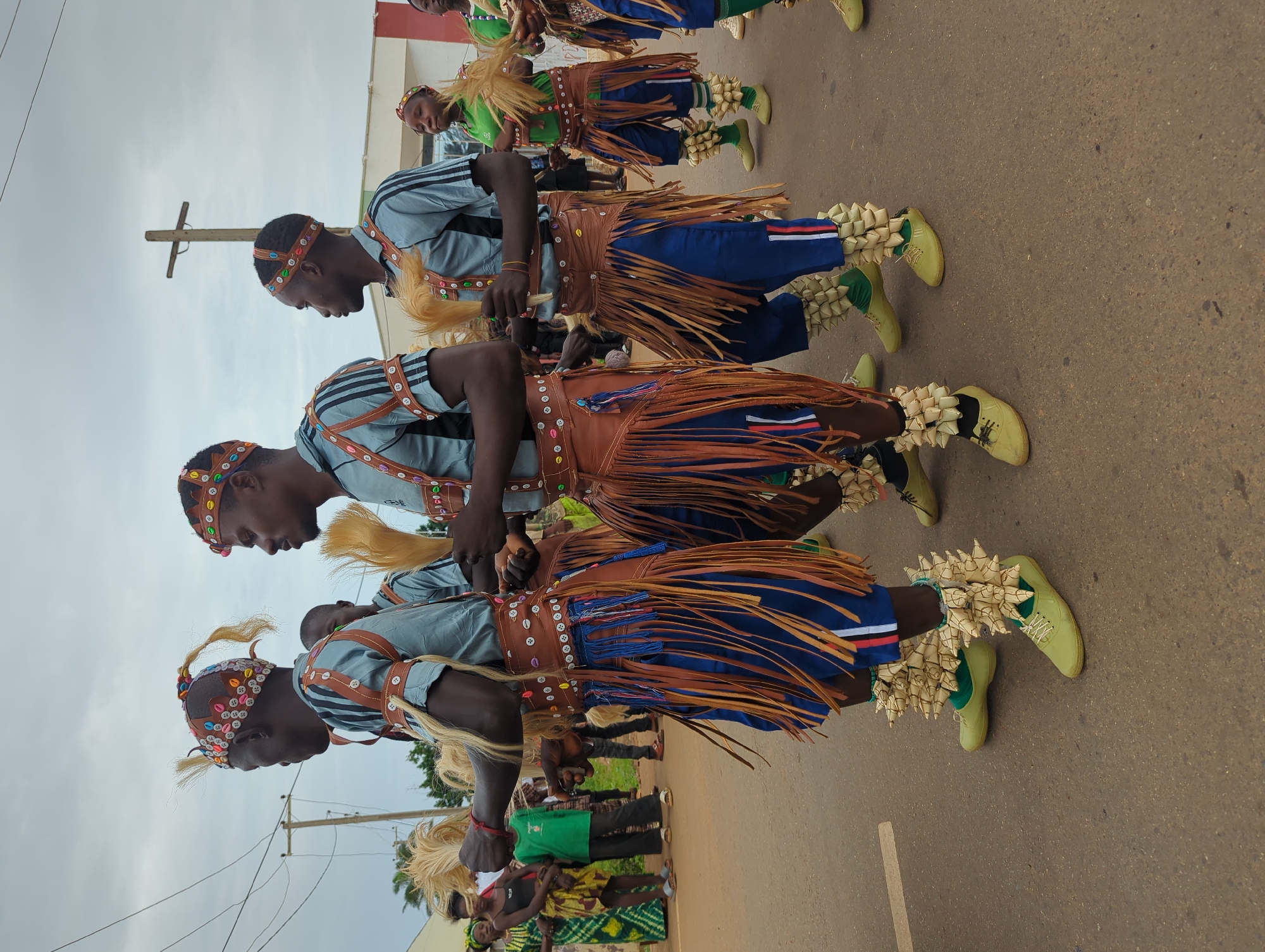 A group of men are performing a dance on a paved surface in what appears to be Maigizo, Nigeria. They are dressed in traditional clothing, including blue shirts with striped trim, brown leather harnesses and skirts with fringe, and light green shoes and socks. Decorations of tan woven or straw material are attached to their ankles and their headwear. The men are lined up in a row, facing the same direction, and appear to be moving in unison. Other people are visible in the background, watching the performance. A wooden cross is visible in the background. Power lines are visible in the sky.