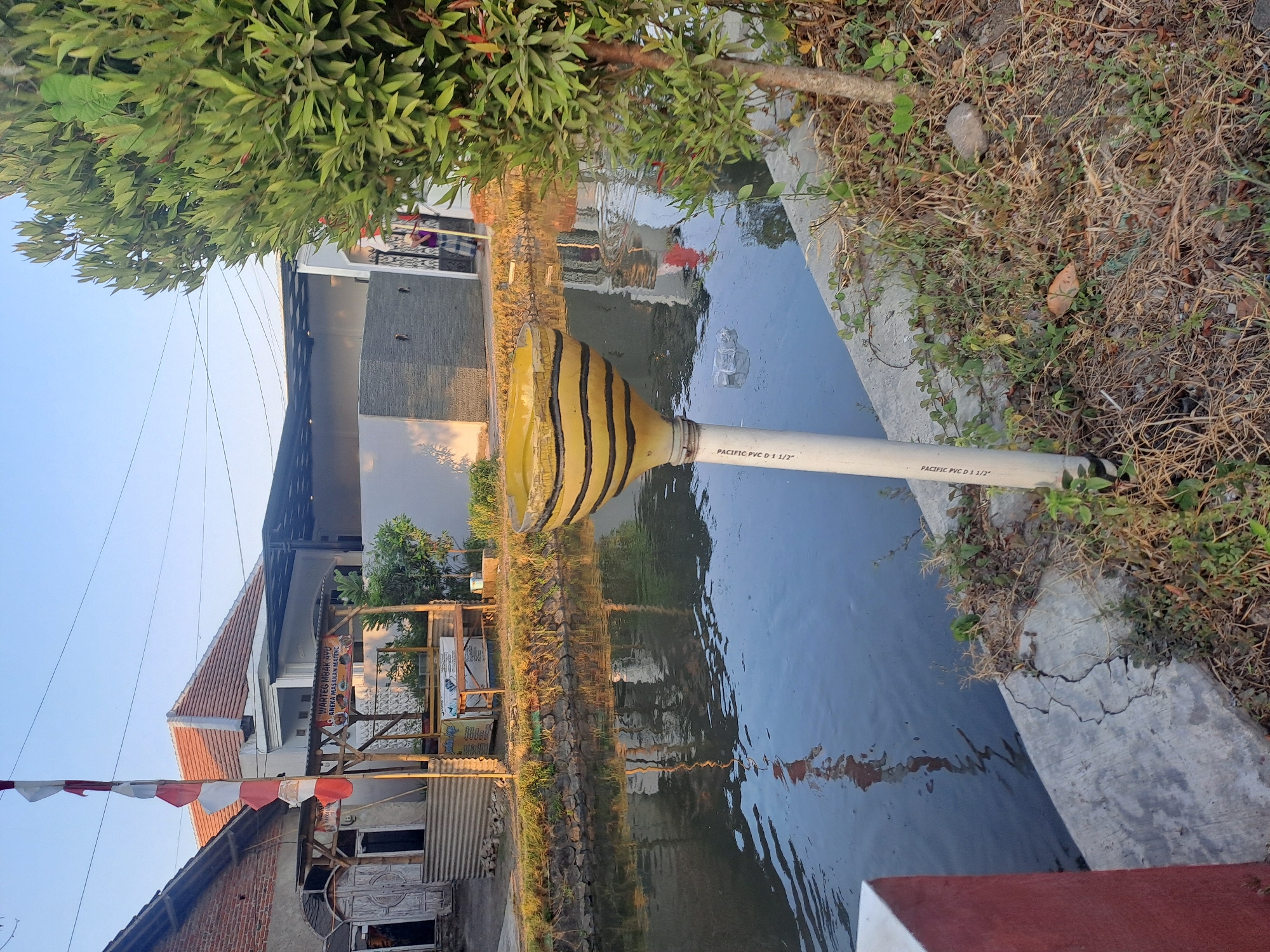 The image depicts a narrow canal with calm, reflective water, situated in what appears to be a residential or semi-rural area. On the left bank of the canal, a white concrete embankment is visible, with a red and white striped flag pole and a building with a reddish-brown brick exterior and a corrugated metal wall. Adjacent to this is a small wooden structure with a thatched roof, serving as a food stall or warung, indicated by signs that read "WARTEG MBAK AYU" and "ANEKA MASAKAN BERSENSUNG." Beyond the warung, a modern house with a light-colored facade and a dark, sloped roof is present.
On the right bank of the canal, a large, dense green tree with reddish accents on its leaves dominates the scene. In the foreground, a white PVC pipe is erected, topped with a yellow and black striped decorative funnel or planter. The ground on this side is covered with dry grass, weeds, and some scattered stones. The overall lighting suggests it is either early morning or late afternoon, with soft, warm light casting shadows. The presence of the warung and residential buildings points to a community setting. The canal itself appears to be part of the urban or suburban infrastructure. The environment is clean with no visible litter except for a few plastic items near the white pipe in the canal.