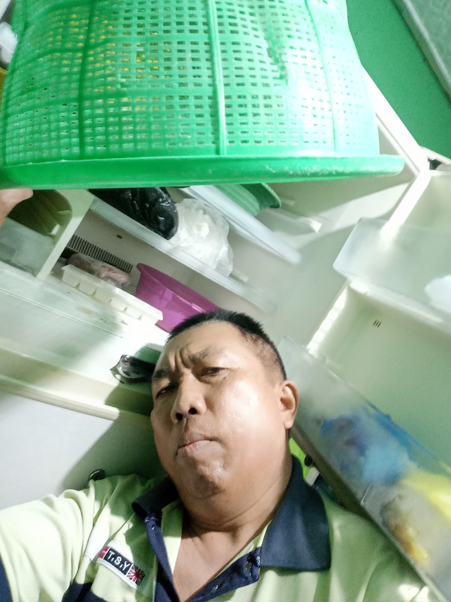 A middle-aged East Asian man, wearing a light green polo shirt with a navy collar and a patch that reads "T.S.Y.", is taking a selfie inside an open refrigerator. He has short black hair, dark eyes, and a serious expression. The refrigerator is mostly empty, with a few items visible on the shelves. These include a green plastic basket, a black trash bag, white plastic bags, an ice cube tray, and a purple basin. The lighting is dim and appears to be from the refrigerator's interior light. The scene appears to be indoors, but the specific location within a city is not determinable from the image.