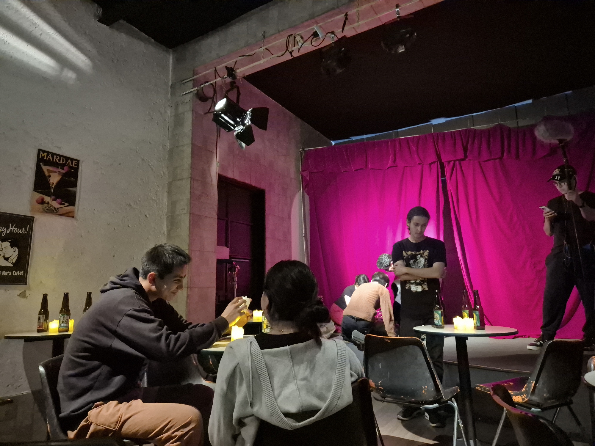 This image depicts an indoor scene, likely a small theater, bar, or rehearsal space, possibly in Puebla, Mexico. The setting features a performance area with a vibrant pink curtain backdrop and an audience section with tables and chairs.
On the left, a man in a dark hoodie and brown pants is seated at a table with a woman, whose back is to the camera, wearing a grey hoodie. The man has white smudges on his face, possibly makeup, and is looking down, holding what appears to be a glowing object, possibly a phone or a candle. On their table and another nearby, several brown and green beer bottles are visible, along with small, lit electric candles or lamps. One green bottle clearly shows the brand "INDIO".
On the back wall, two posters are visible. The top one reads "MARDAE" above an illustration of a martini glass. Below it, part of another poster shows "py Hour!" and "He's Cute!", suggesting "Happy Hour!"
The stage area is illuminated by several spotlights mounted above. On the stage, two individuals are seen in a kneeling or bending position, appearing to be arranging something or rehearsing. Standing to their right, a man in a black t-shirt with a graphic design stands with his arms crossed, observing the activity. Further to the right, a person in a black cap and t-shirt holds a boom microphone, looking at a phone, indicating a recording or filming session is in progress.
The walls are light-colored and have a rough, textured finish. The overall ambiance is casual, suggesting a creative project, rehearsal, or small performance event occurring in the evening or at night, given the artificial lighting.