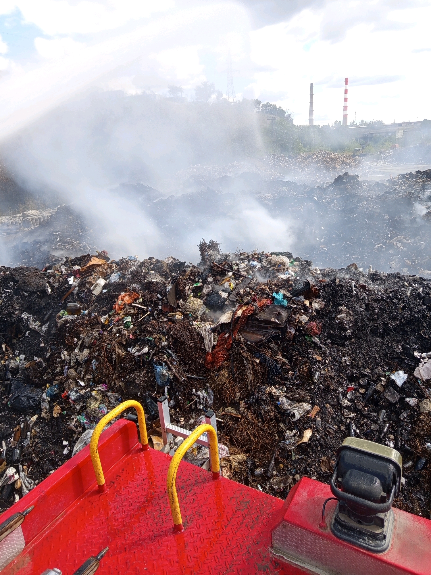 The image shows a landfill fire in Tiraspol, Moldova. Thick white smoke billows from a vast pile of smoldering trash, which appears to be a mix of household waste, plastic, and other debris. In the foreground, a red vehicle with yellow railings is visible, suggesting a firefighting or cleanup operation is underway. In the background, industrial chimneys, one with red and white stripes, rise above the smoke and a dense line of trees. The sky is overcast with clouds. The scene depicts an environmental hazard and ongoing emergency response.