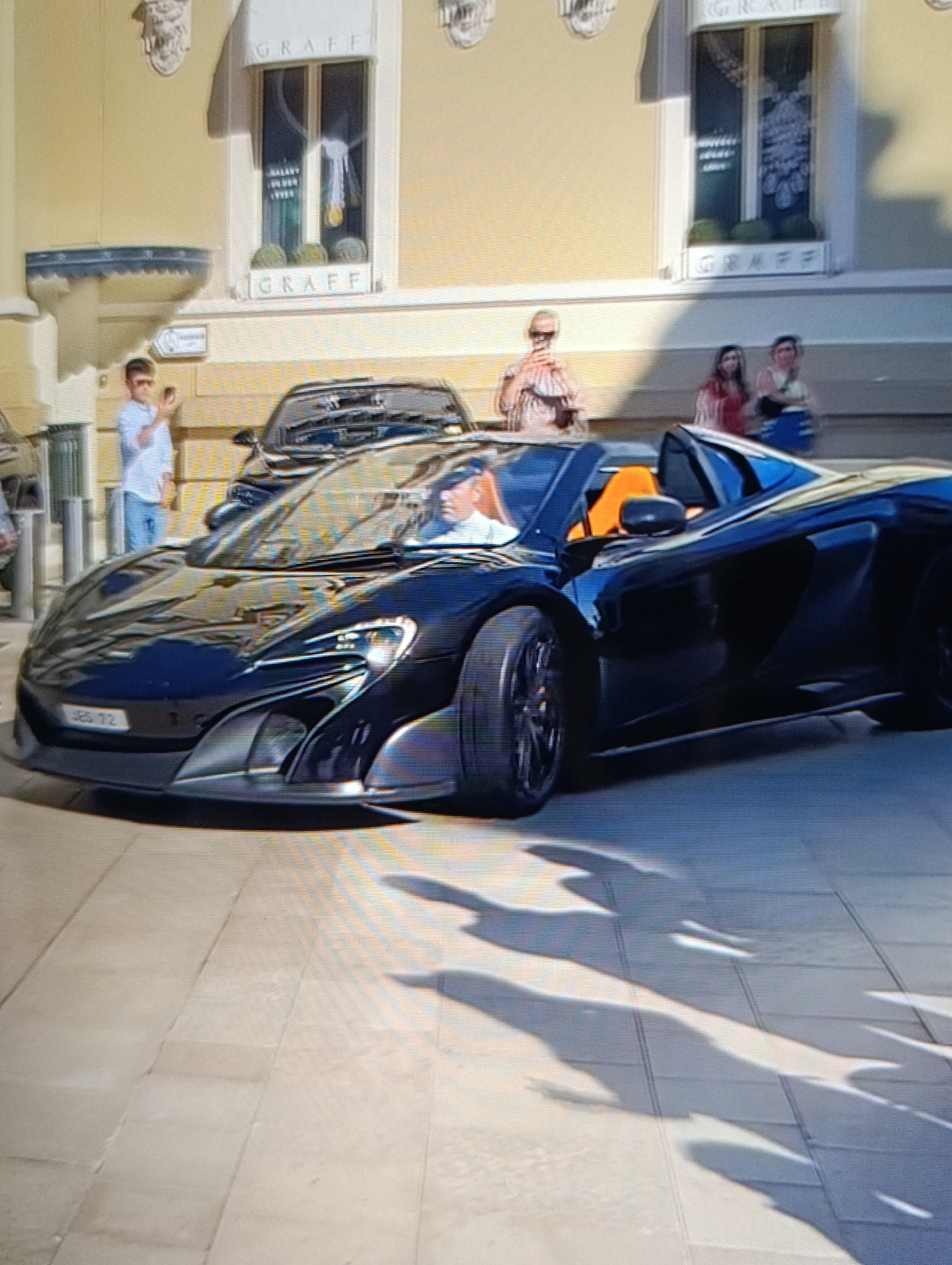The image shows a dark blue McLaren 675LT Spider parked on a light-colored tiled surface. The car has an orange interior, visible in the open cabin. A man is sitting inside the car. Behind the McLaren is another car, and several people are standing on the sidewalk. One person is taking a picture with their phone, while others are observing the scene. Above the people, there are windows with "GRAFF" signage. The location is in the city of Unknown, Unknown.