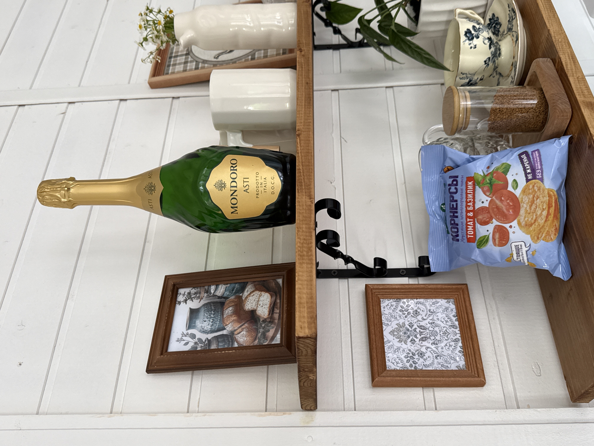 A close-up, eye-level shot shows a wooden shelf with various items, likely in a kitchen or dining area in Starokorsunskaya, Russia. The shelf is made of unfinished wood and is supported by black metal brackets.
On the shelf, from left to right, are:
* A small, framed picture with a wooden frame, displaying a decorative pattern.
* A large green bottle of Mondoro Asti sparkling wine with a gold foil cap.
* A white ceramic mug.
* A white ceramic pitcher containing a small bouquet of white flowers with yellow centers.
* A framed picture with a wooden frame, showing a still life of bread and pottery.
* To the right of the Mondoro bottle, partially obscured, is a framed picture with a plaid pattern.
Below this shelf, on a lower wooden shelf, are:
* A blue bag of "Korneri" tomato and basil flavored crisps.
* A glass jar with a wooden lid filled with small brown seeds.
* A teacup and saucer with a blue floral pattern.
* A green potted plant.
The background is a white, horizontally paneled wall. The lighting is natural and bright, suggesting daytime. There are no people or animals visible in the image.