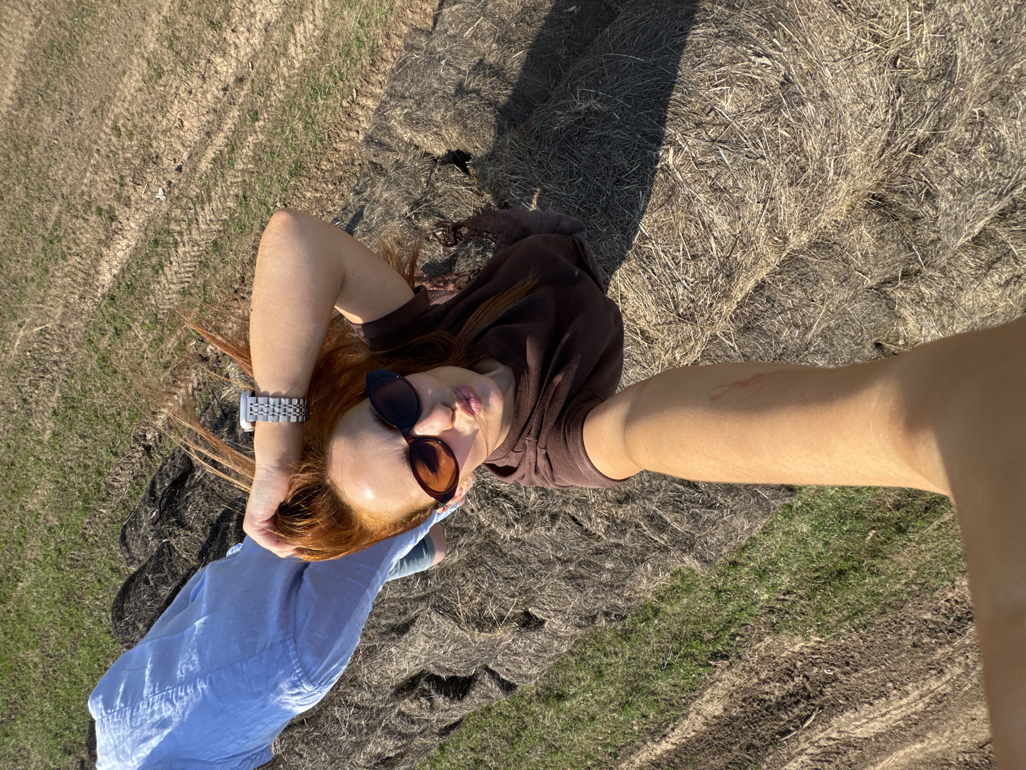 A person with reddish-orange hair and wearing sunglasses is lying on a surface covered with dried grass or hay. The person is wearing a brown shirt and a light blue garment. A silver watch is visible on their wrist. The scene appears to be in a rural environment. The location is Sivinskoe, Russia.