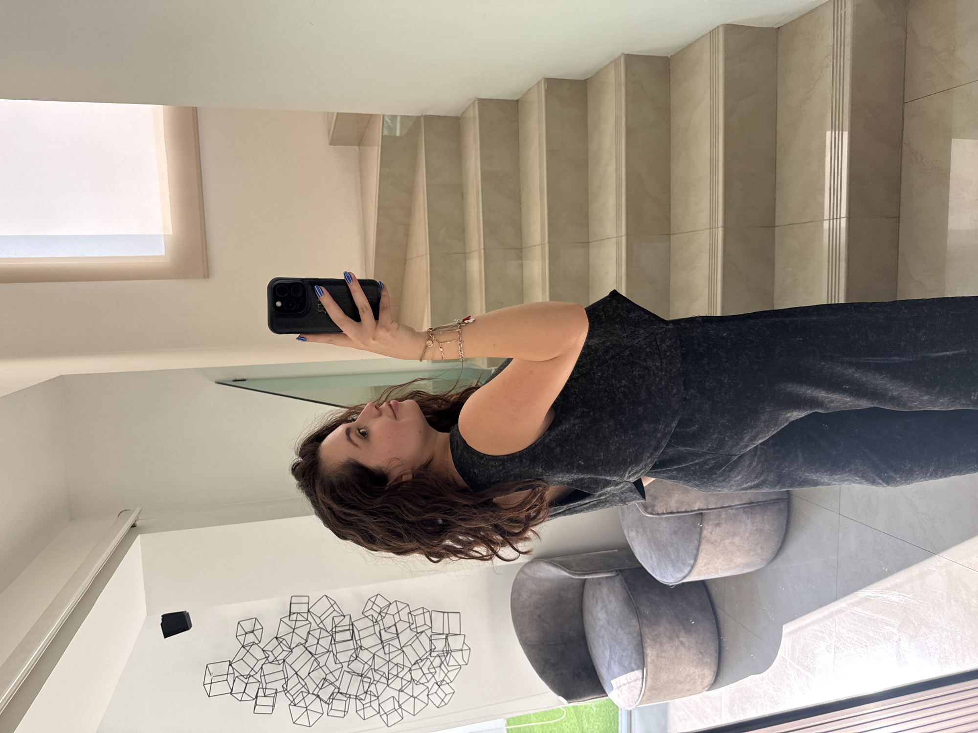 A woman is taking a selfie in what appears to be a modern, brightly lit home. She is holding a black smartphone with blue nail polish on her fingernails. The woman has long, wavy brown hair and is wearing a gray outfit. Behind her are two modern gray chairs and a black geometric wall sculpture. A staircase with light-colored steps is visible, along with a large window. The location is likely a high-end residence. The city of Santa Clara Ocoyucan, Mexico, has not been specifically identified in the image.