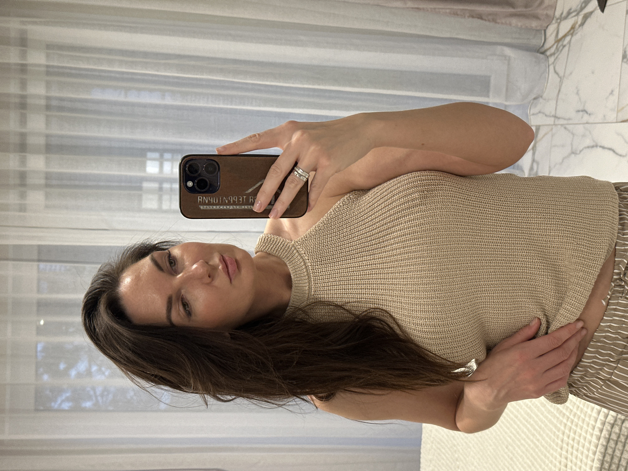A woman with long brown hair is taking a selfie indoors. She is holding a phone with a brown wooden case in her right hand, displaying the back of the phone to the viewer. Her left hand is near her waist. She is wearing a light tan knitted top and beige and white striped bottoms. A ring is visible on her right ring finger. The background features a window with white sheer curtains. The scene is indoors, with a marble-tiled wall partially visible on the right side. There is no evidence to indicate this scene is in Chaikovsky, Russia.