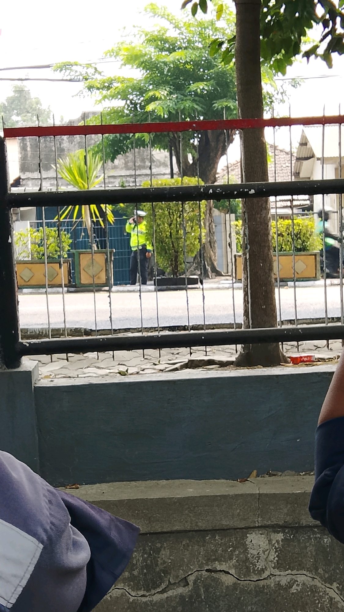 A police officer in a neon yellow vest and dark pants stands on the side of a paved road, holding a white object to his ear, possibly a radio or phone, in Gapurosukolilo, Indonesia. He is positioned behind a black metal fence with red horizontal accents. Behind the officer, there are planters with greenery, trees, and buildings. In the foreground, a dark grey concrete structure is visible, with a portion of a dark blue garment with a white patch on the left side.