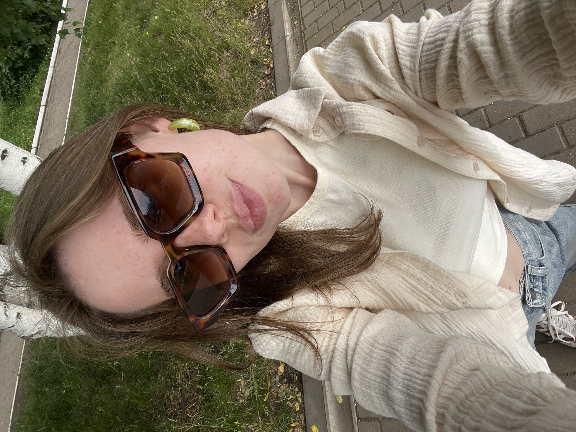 A young woman is taking a selfie outdoors. She is wearing a cream-colored, textured button-up shirt over a cream-colored crop top, light wash denim jeans, and white sneakers with red accents. She has on large, brown tortoiseshell sunglasses and a single, large, light green hoop earring in her right ear. Her lips are slightly puckered. The background shows a grassy area with a birch tree and a paved walkway. The scene suggests a casual outdoor setting in a city park or on a sidewalk. This image was taken in Chaikovsky, Russia.