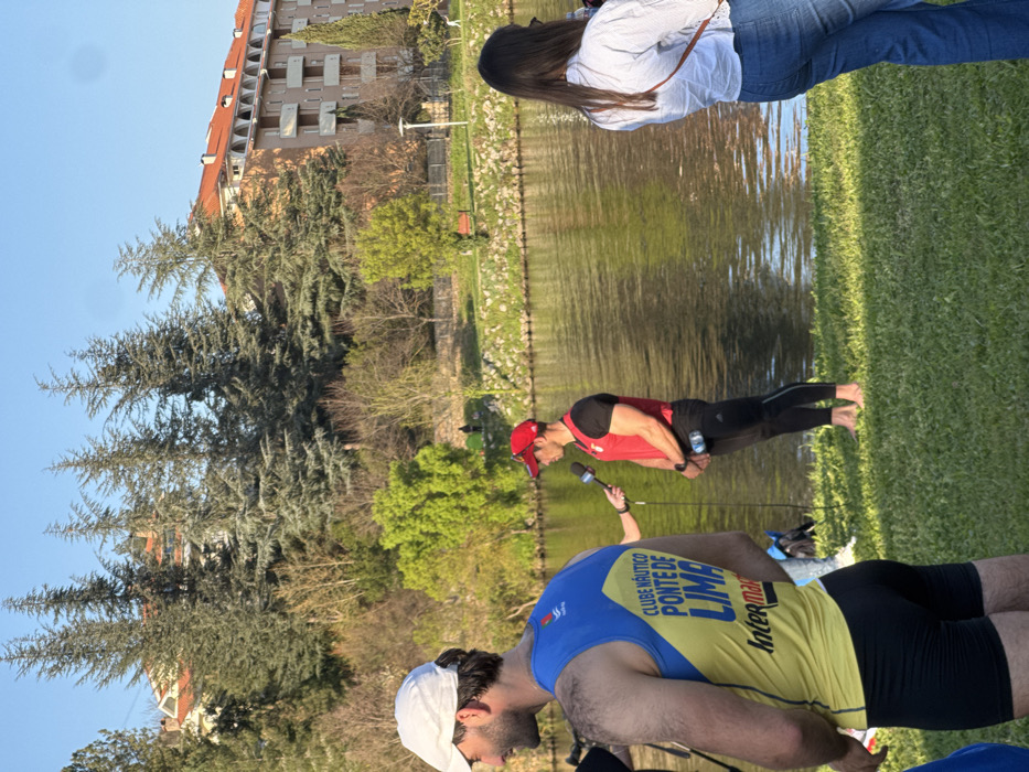 The scene depicts an outdoor interview by a body of water in Mirandela, Portugal, under clear blue skies with strong sunlight. In the mid-ground, a male subject stands barefoot on green grass, facing left, wearing a red baseball cap, a red sleeveless athletic top, and black athletic leggings. He holds a small water bottle in his left hand. An unseen individual extends a microphone with a black foam cover and a white logo, possibly "TVI," towards him for an interview.
To the immediate left of the interviewed subject, partially in the foreground, the back of another male subject is visible. He wears a white baseball cap, a yellow and blue sleeveless athletic vest with the text "CLUBE NÁUTICO PONTE DE LIMA" and "Intermarché," and black athletic shorts. To the right of the interviewed subject, also with her back to the camera, stands a female with long dark hair, dressed in a light-colored long-sleeved collared shirt and blue jeans, with a tan strap across her body.
The background features a calm body of water, likely a river or canal, reflecting the sky and surrounding trees. Beyond the water, the far bank is lined with green grass, various trees including tall conifers and deciduous trees with some bare branches and fresh green foliage. Behind the trees, several multi-story residential buildings with terracotta-tiled roofs are visible, along with a prominent light-brown building featuring balconies. A red park bench and a low fence are also discernible on the far bank. A distant, dark human-like figure is faintly visible near a patch of bright green trees on the far bank.