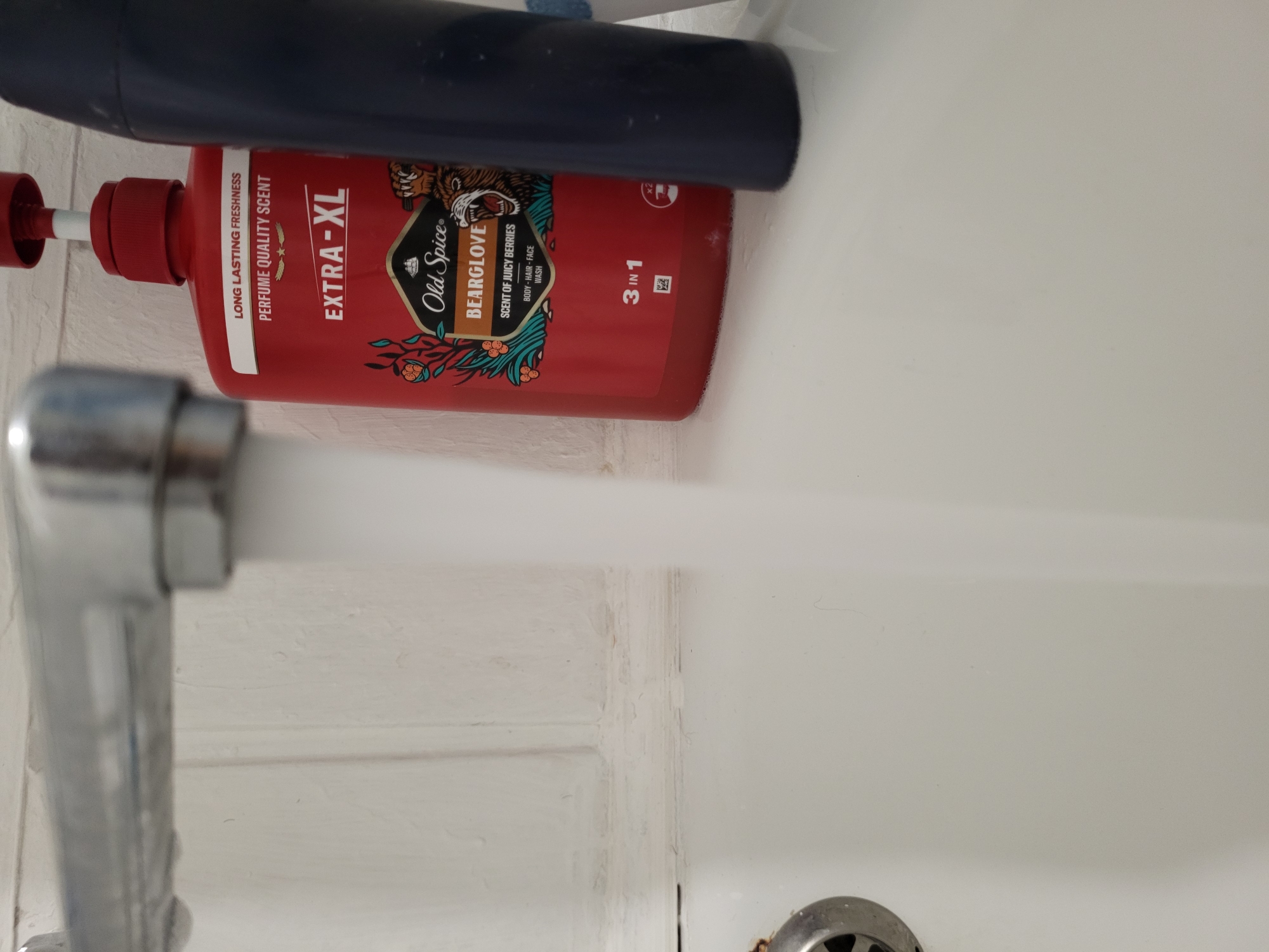 A close-up, eye-level shot shows a silver faucet with water running from it, creating a blurred stream. To the right, a red pump bottle of "Old Spice Bearglove" body wash with "Extra-XL" branding is visible. The bottle has an illustration of a bear's head and berry branches. Next to it is a dark blue bottle. The background consists of a white, textured wall with vertical lines. The foreground is a white sink or bathtub. The scene is indoors, likely a bathroom. No specific location in Horishni Plavni, Ukraine is identifiable from the image.