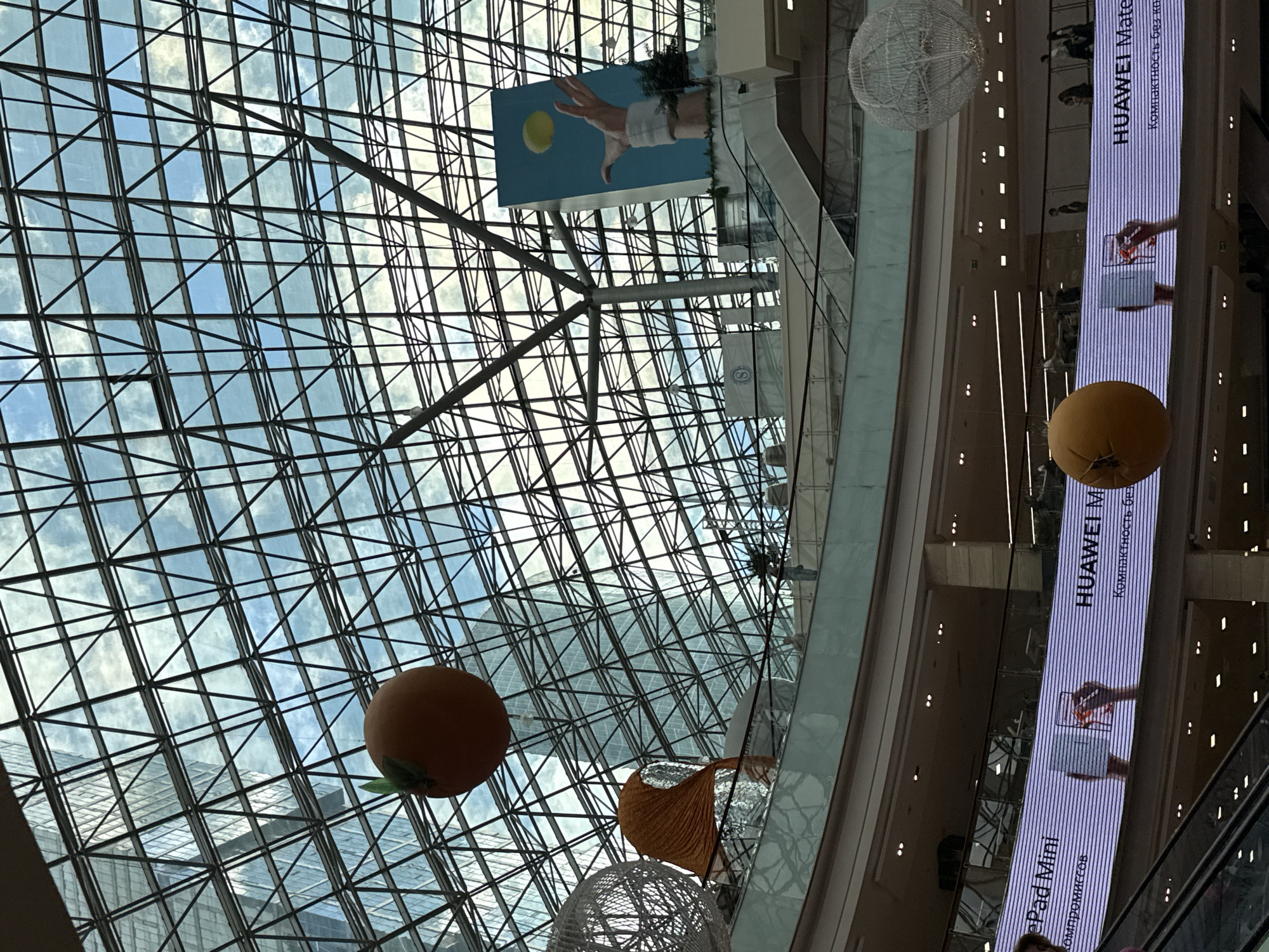 The image captures the interior of the MEGA Khimki shopping mall in Khimki, Russia. The focal point is the geometric grid of the glass roof, partially revealing a bright, cloudy sky. Hanging from the ceiling are decorative elements including spherical orange and white paper lanterns. A large advertisement panel for Huawei products, displaying the text "HUAWEI Mate Compactness без компромиссов" and an image of a hand holding a device, is positioned vertically along the right side of the image. An escalator can be partially seen near the Huawei advertisement panel. Additionally, a banner depicting a hand reaching for a sun-like sphere is visible towards the upper portion of the image.