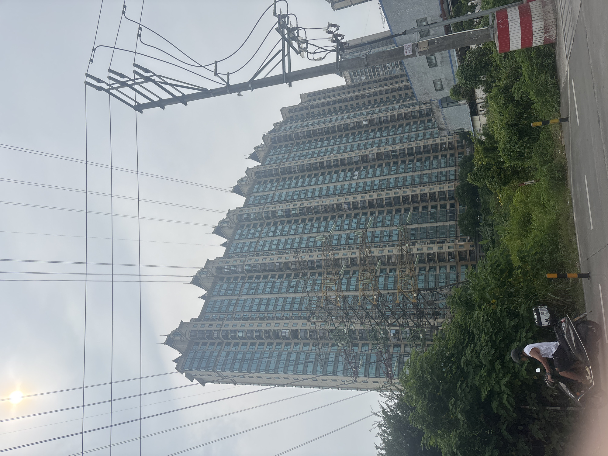 A person wearing a white tank top and black shorts rides a black scooter on a paved road in Nanhai District, China. The scooter has a black storage box on the back. The person is wearing a black helmet. In the background, a tall, light-colored apartment building with many windows dominates the scene. Numerous power lines crisscross the sky above the building. A large metal electricity tower stands to the right of the building, also covered in wires. Lush green trees and vegetation are visible in the foreground and around the base of the apartment building and electricity tower. A red and white striped cylindrical object is positioned on the side of the road to the right. The sky is overcast and hazy.