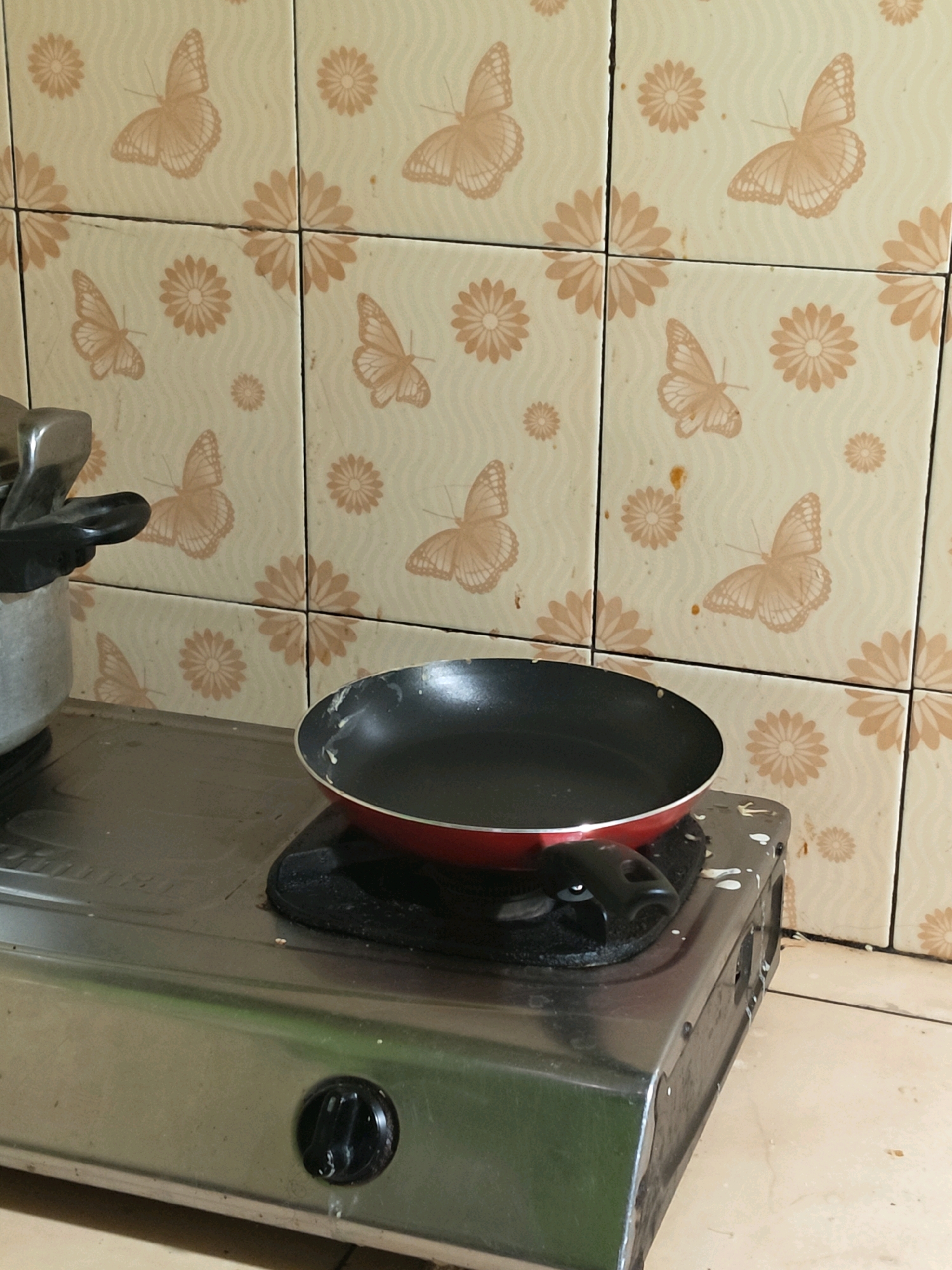 The image shows a close-up view of a gas stove in a kitchen. A black frying pan with a red rim is placed on the stovetop. The stove is located in front of a wall tiled with a pattern of butterflies and flowers. There is a pot to the left of the frying pan, partially visible. The environment appears to be a domestic setting, likely a kitchen in a home. The image does not provide specific details about the city of Sumberwono, Indonesia, beyond its potential location. There are no people visible in the image, and no actions are being performed.