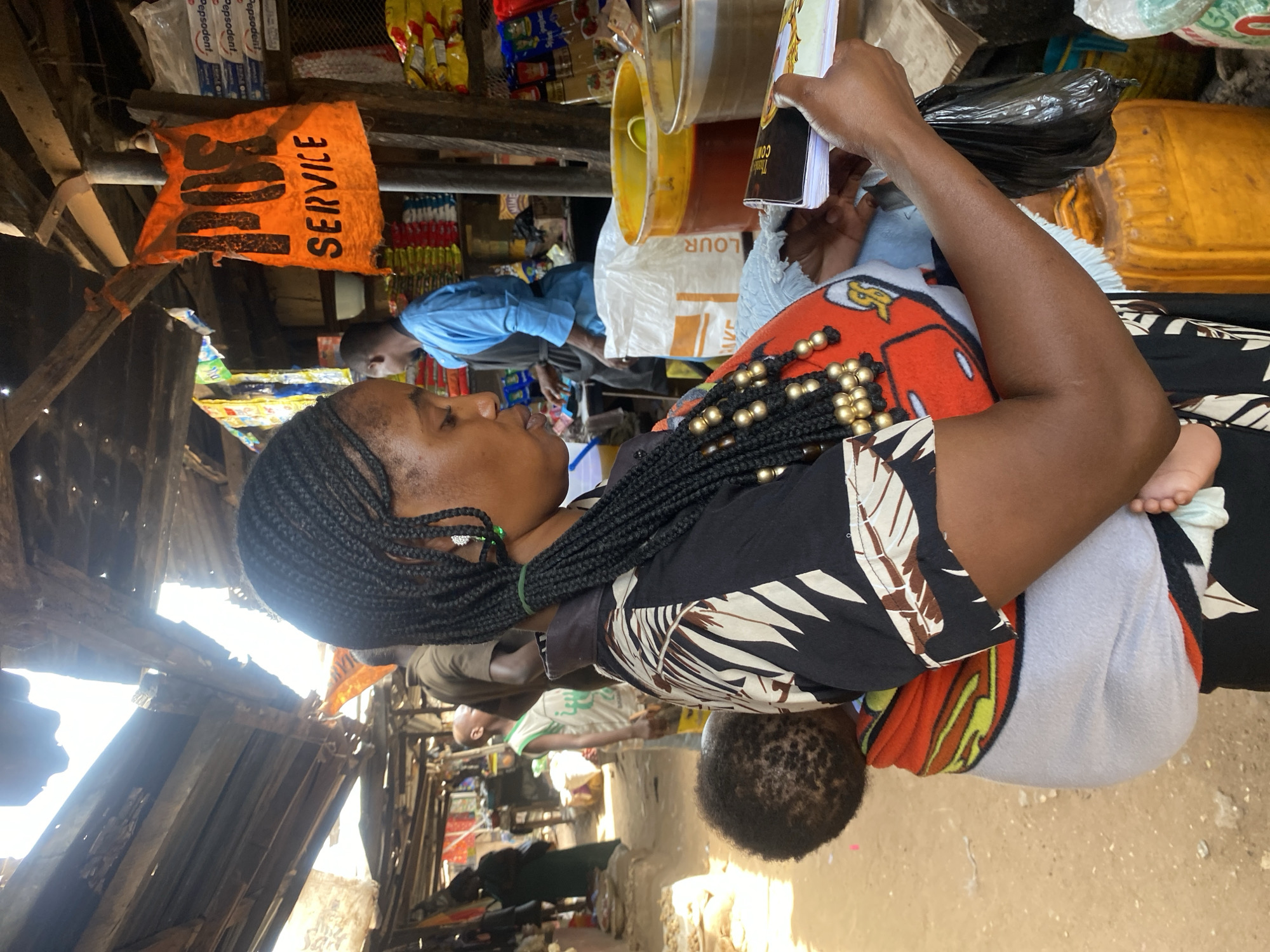 The image depicts a crowded market scene in Rido, Nigeria. In the foreground, a dark-skinned woman is positioned slightly to the left, looking down at objects in her hands. She wears her black hair in tight braids, some adorned with gold beads, and has a small, light-colored earring in her visible ear. Her top features a black and white abstract pattern on the sleeves and shoulders, with an orange, graphic-printed section on the chest. A baby with dark, curly hair is strapped to her back, secured by a patterned orange, yellow, and white cloth, with one small foot visible. The woman holds a small booklet with a dark cover and white pages in her left hand and a black plastic bag in her right.
Behind the woman, a stall or shop setting is evident. A large white bag labeled "FLOUR" is visible on the floor, next to a yellow plastic bucket containing a reddish-brown liquid, likely oil. Further back, shelves display various packaged goods, including stacks of items in red and yellow wrappers, bottles of sauces, and packages of "Pepsodent" toothpaste. An orange banner or piece of fabric with "POS SERVICE" printed in black hangs above these shelves.
In the mid-ground, a man in a light blue long-sleeved shirt and a dark apron stands behind the stall counter, facing away from the viewer. Other individuals are blurred in the background, moving through the market space. The environment features a rustic, wooden structure forming the roof and supports, with corrugated metal sections allowing bright sunlight to penetrate. The ground is a light-colored dirt surface.